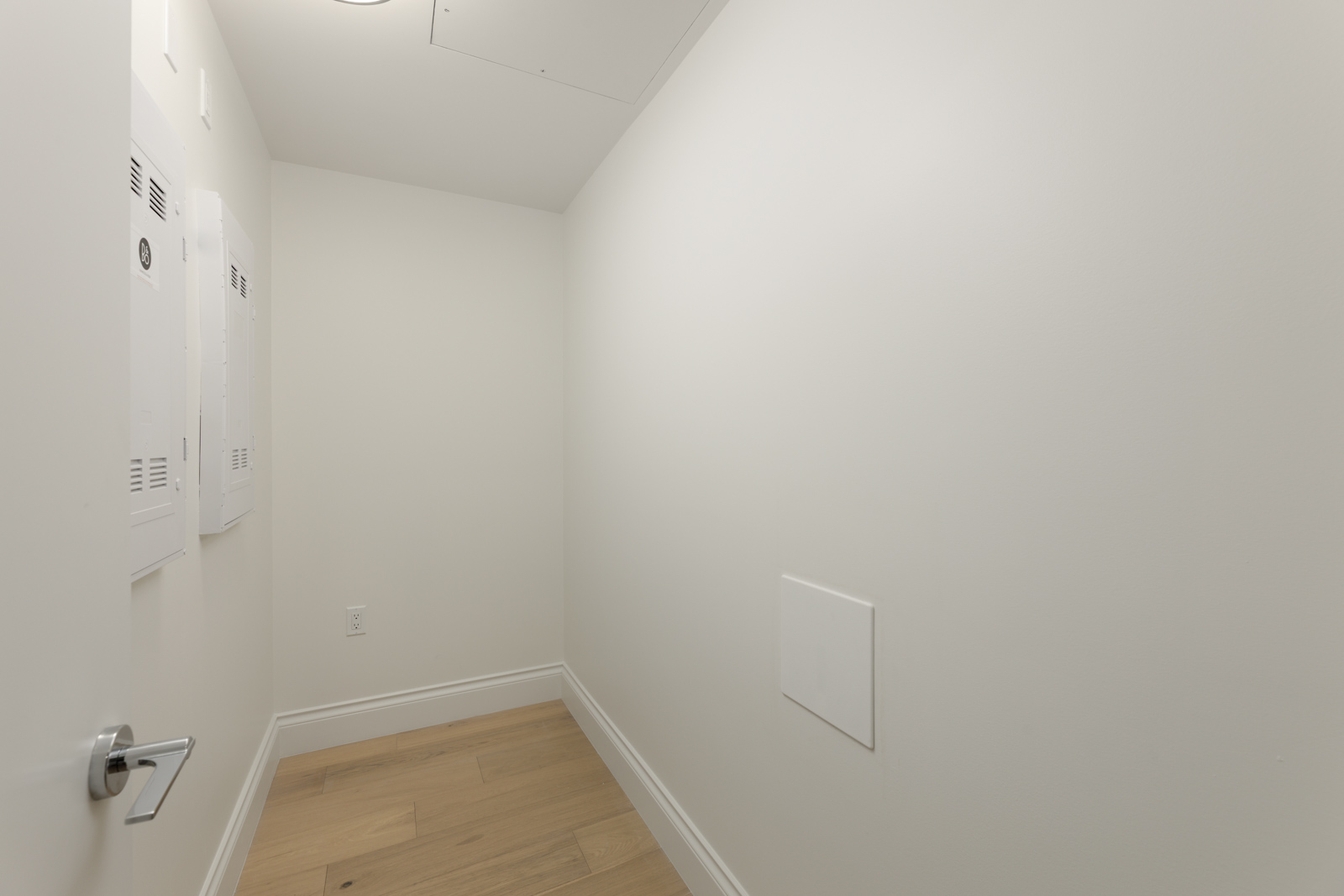 Small, empty white utility room with light wood flooring, access panels on the walls, and a closed door with a silver handle.