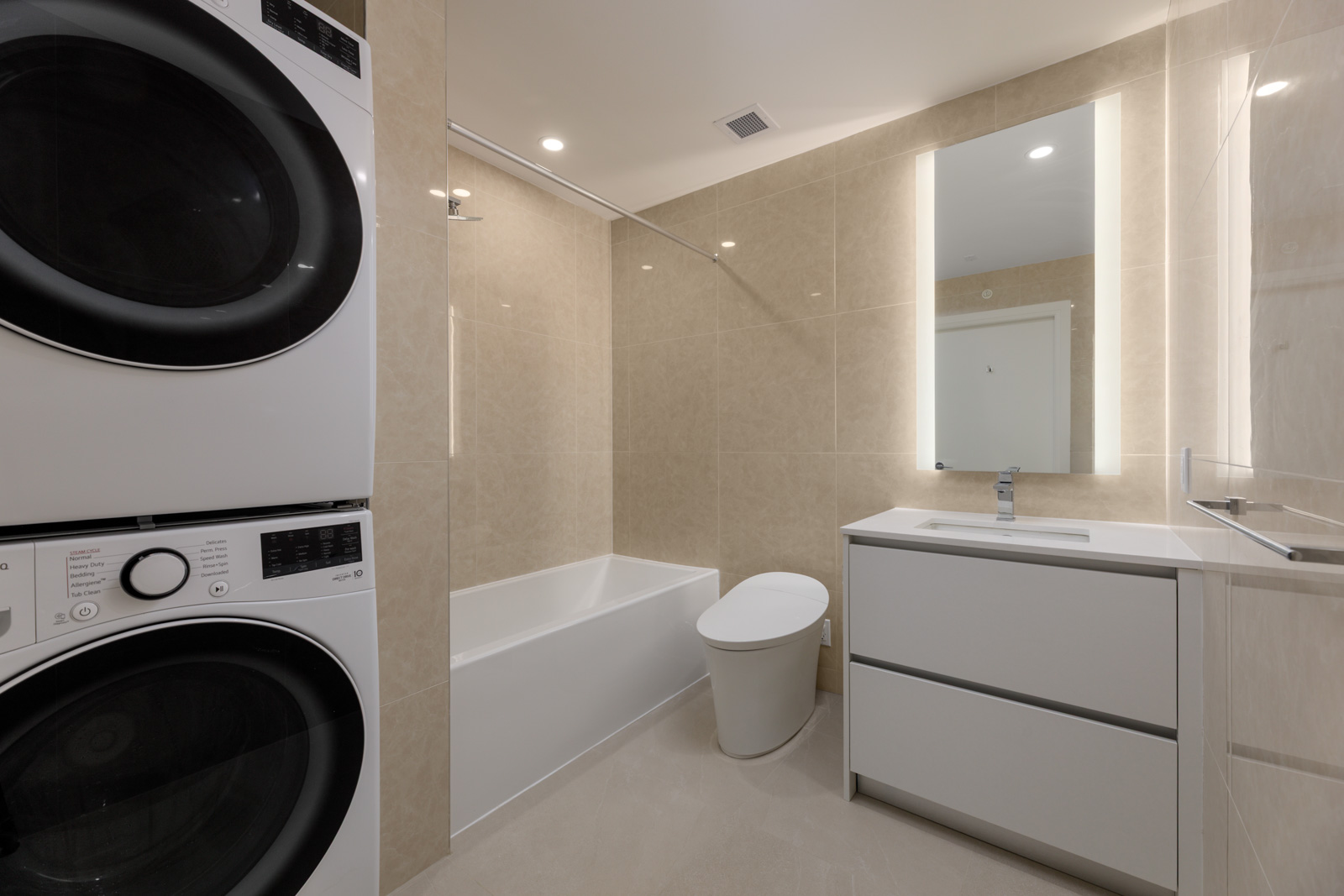 Modern bathroom with beige tiles, a stacked washer and dryer, a bathtub with shower, a white toilet, and a vanity with a mirror and integrated lighting.