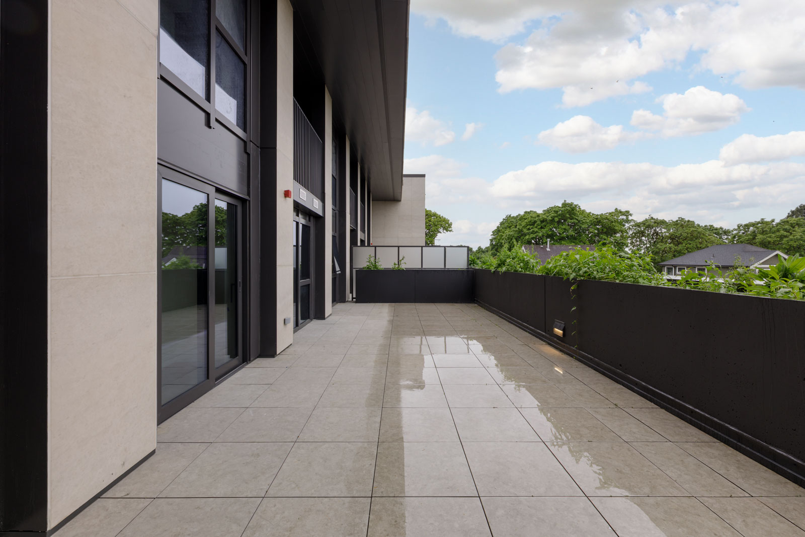 Modern building balcony with large tiled floor, glass doors, black railing, and some water reflecting the sky; trees and cloudy sky visible in the background.