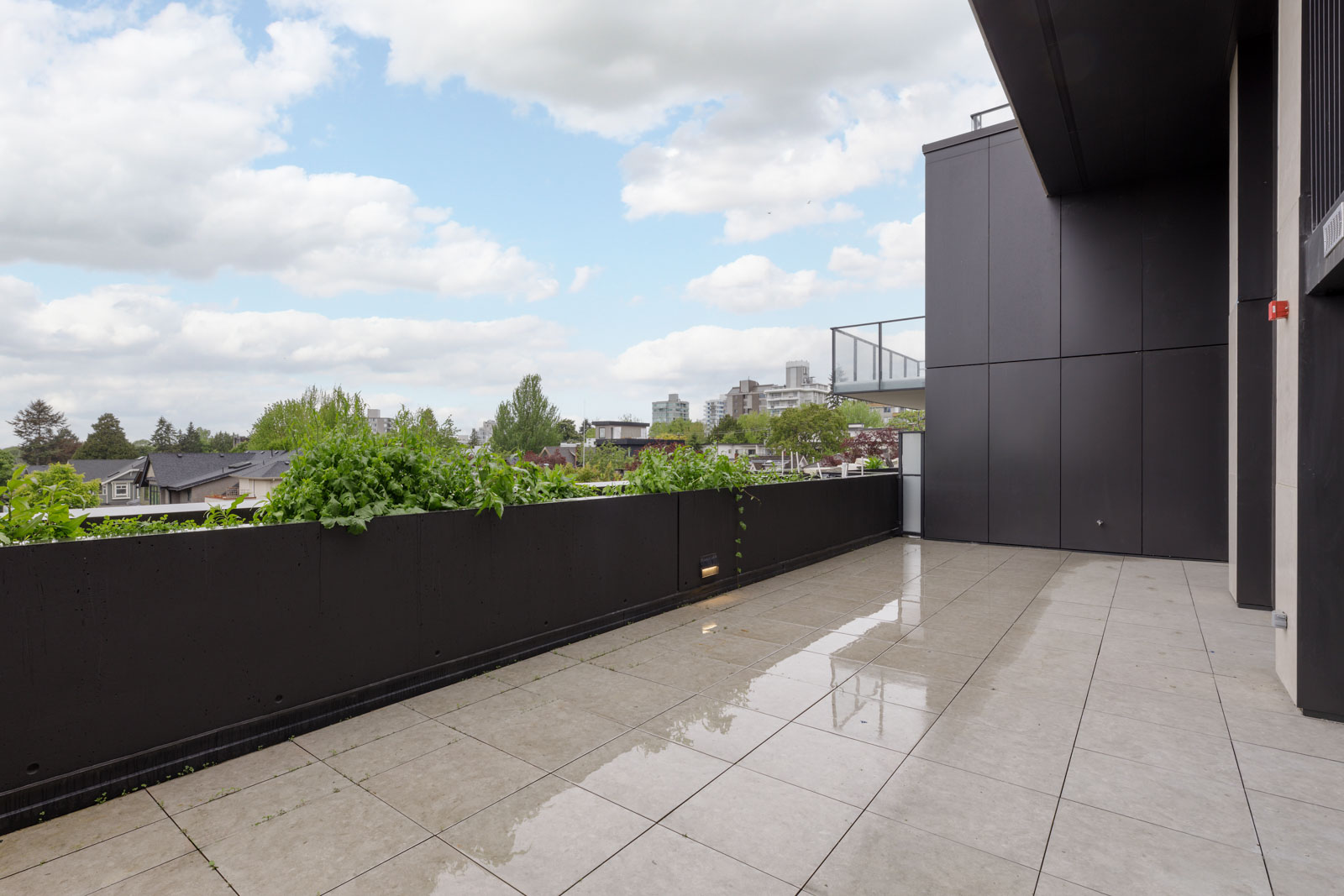 Large outdoor terrace with wet tiled floor, black walls, and a perimeter planter, overlooking rooftops and trees under a partly cloudy sky.