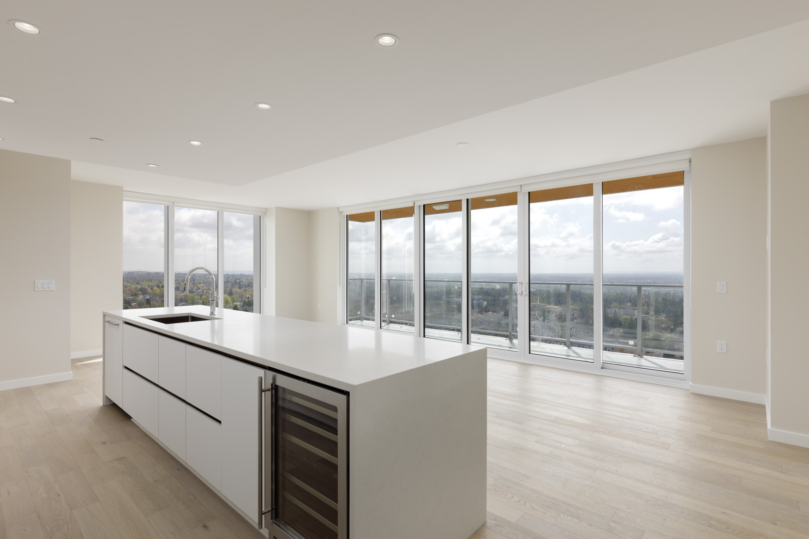 A modern, empty apartment with light wood floors, a large kitchen island, floor-to-ceiling windows, and a balcony overlooking a cityscape.