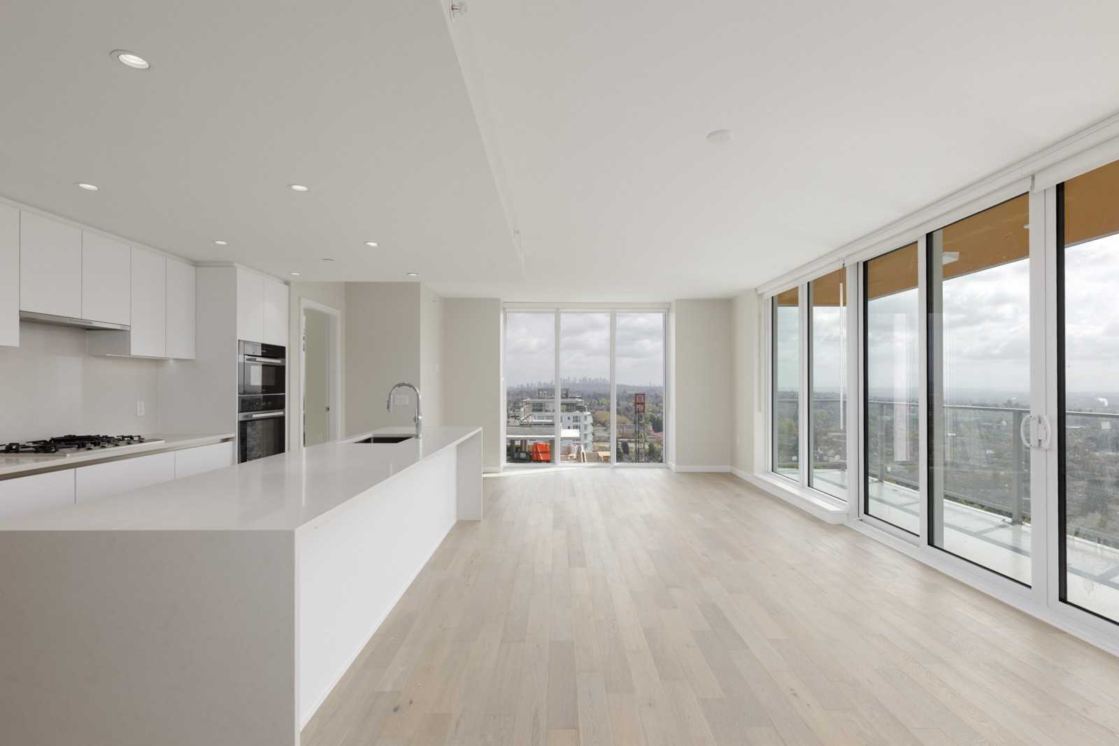 Spacious modern apartment with light wood floors, large windows, open kitchen with white cabinets, and a city view from the balcony.