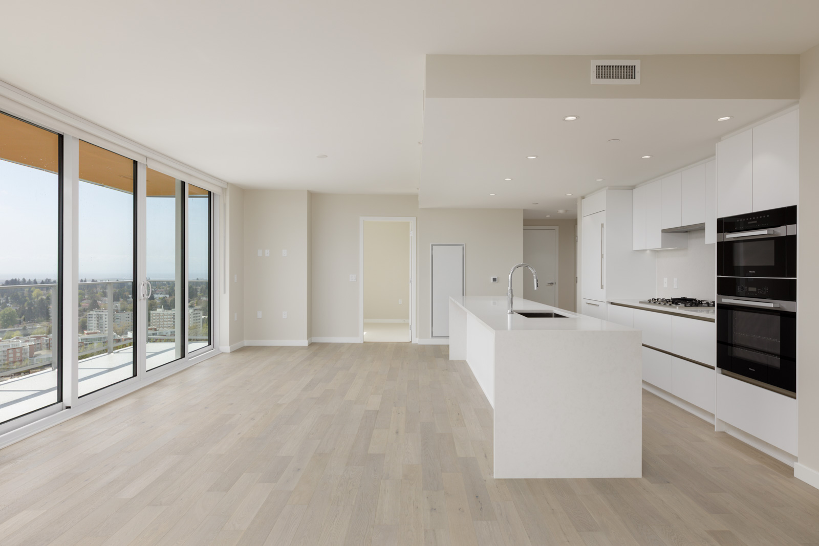 Modern, unfurnished apartment featuring an open-plan kitchen with white cabinetry, built-in appliances, light wood floors, and large windows offering a city view.