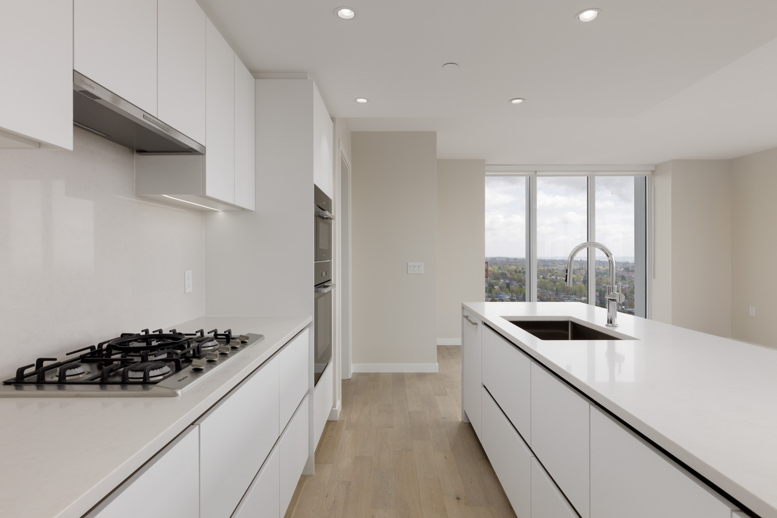 Modern kitchen with white cabinets, gas stove, built-in ovens, large island with sink, and floor-to-ceiling windows overlooking a cityscape.