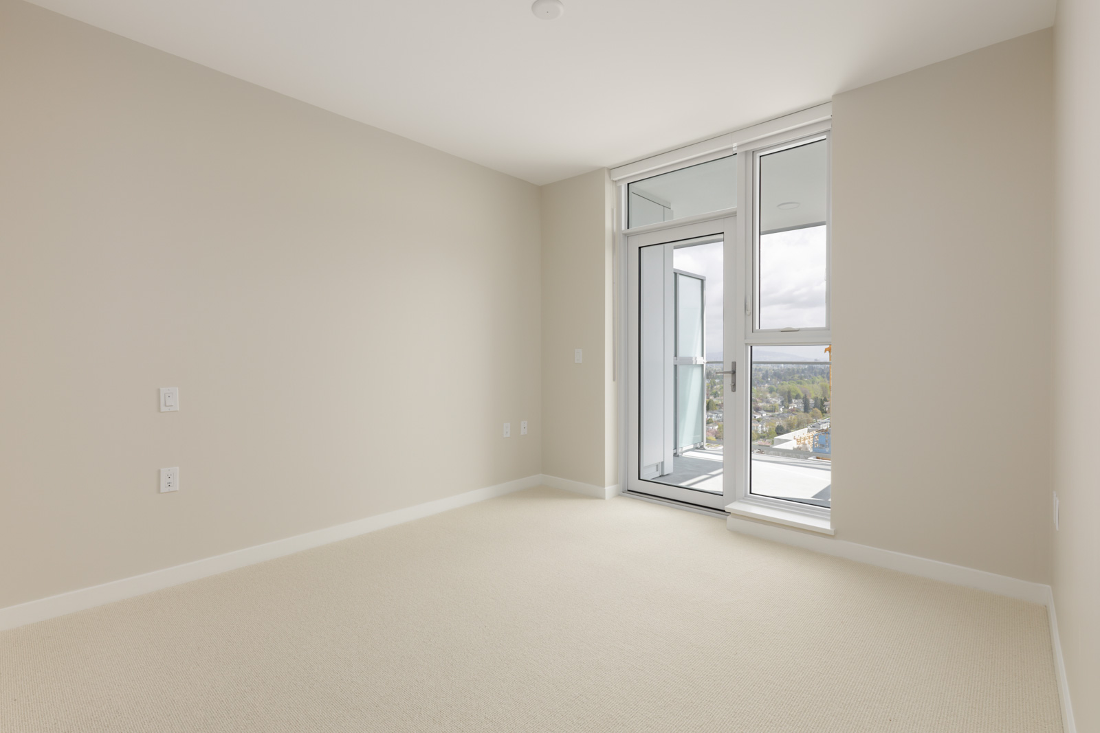 Empty room with light-colored carpet and beige walls, featuring floor-to-ceiling windows and a glass door leading to a balcony.