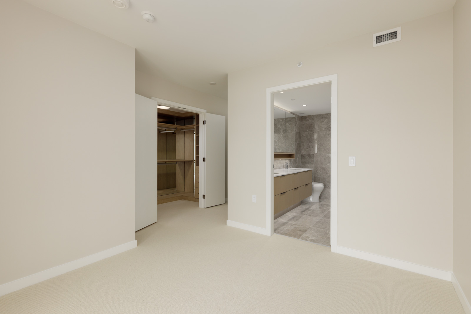 A beige carpeted bedroom with an open door to a walk-in closet on the left and an en suite bathroom with gray tile and double vanity on the right.