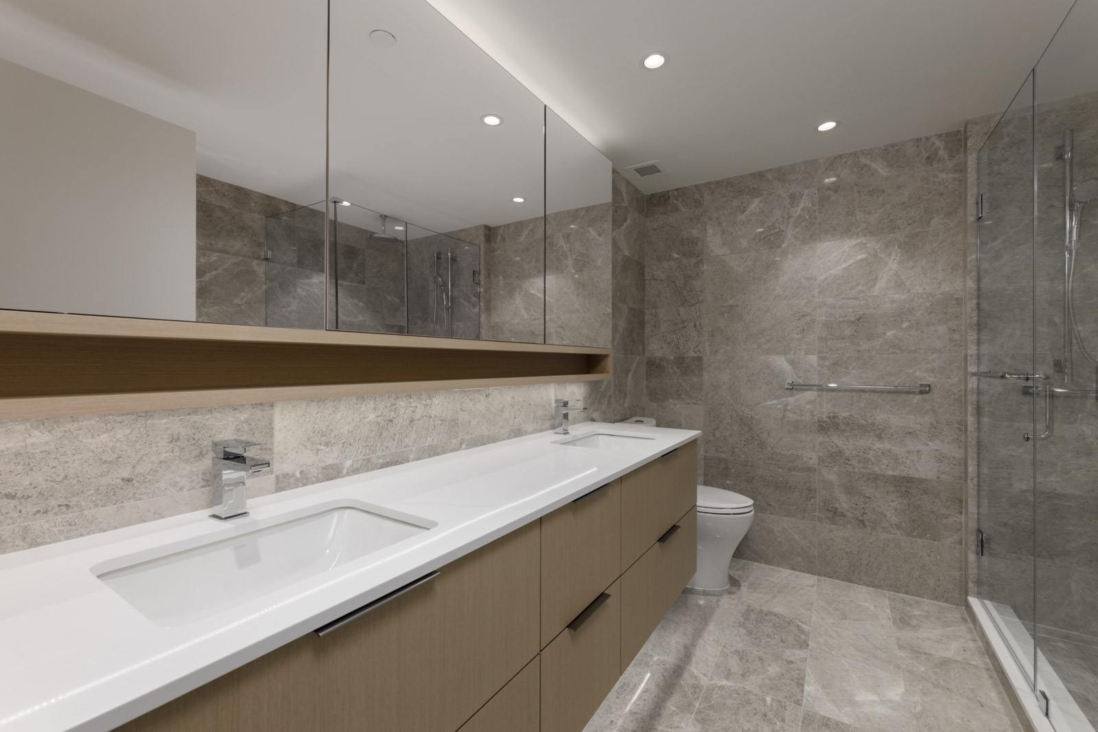 Modern bathroom with double sinks, large mirrors, wood cabinetry, marble walls, floor tiles, a toilet, and a glass-enclosed shower.