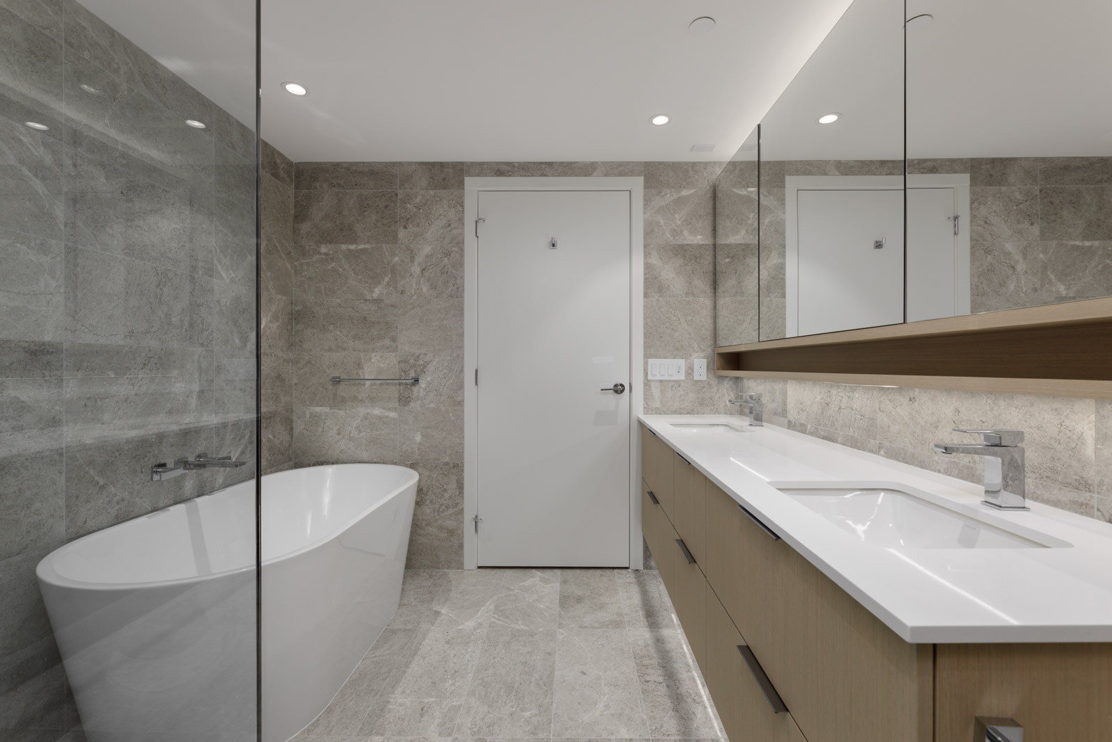 Modern bathroom with gray marble walls and floor, a white freestanding bathtub, a double sink vanity with wood cabinets, and a large mirror above.