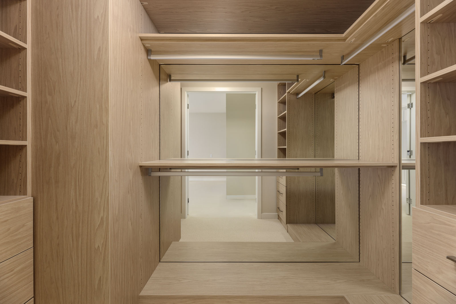 Spacious walk-in closet with wooden shelves, drawers, and hanging rods, featuring a large mirror and open doorway to an adjacent room.