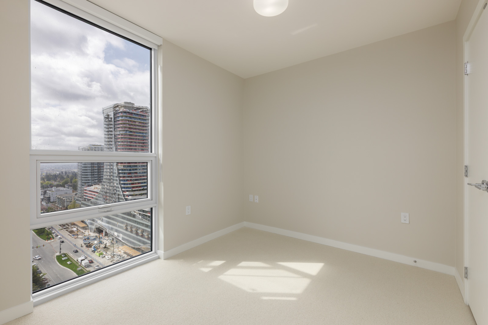 Empty beige-walled room with a large window showing city buildings and streets outside; white ceiling, closed door, and light fixture visible.