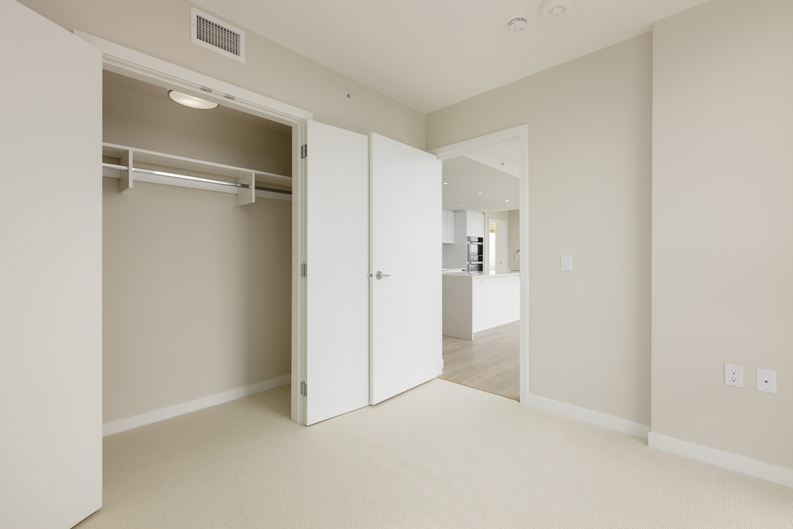 Empty beige-walled bedroom with open closet and door, carpeted floor, and view into a modern white kitchen area.