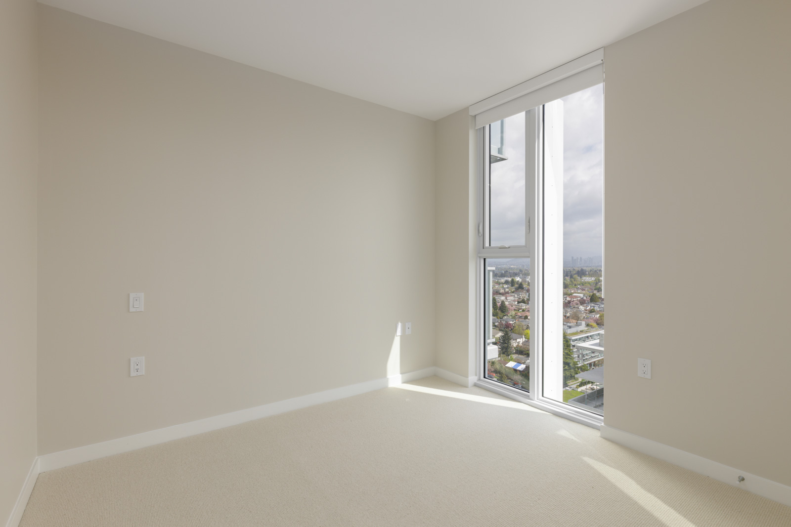 Empty room with beige walls and carpet, a large window on the right side letting in natural light and showing a city view outside.