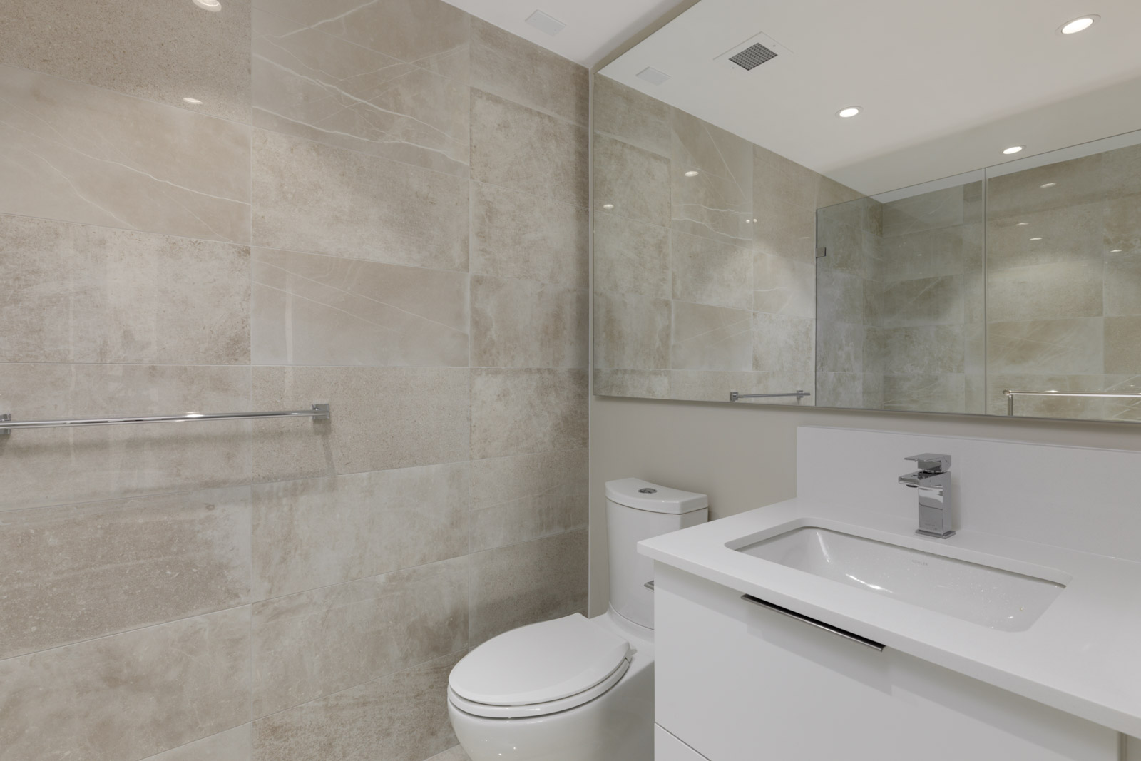 Modern bathroom with beige tiled walls, a white toilet, a white vanity with integrated sink, a chrome faucet, and a large wall mirror.