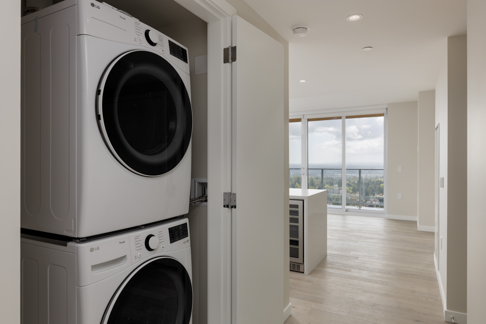 Stacked washer and dryer in a closet next to an open living area with large windows and light wood flooring.