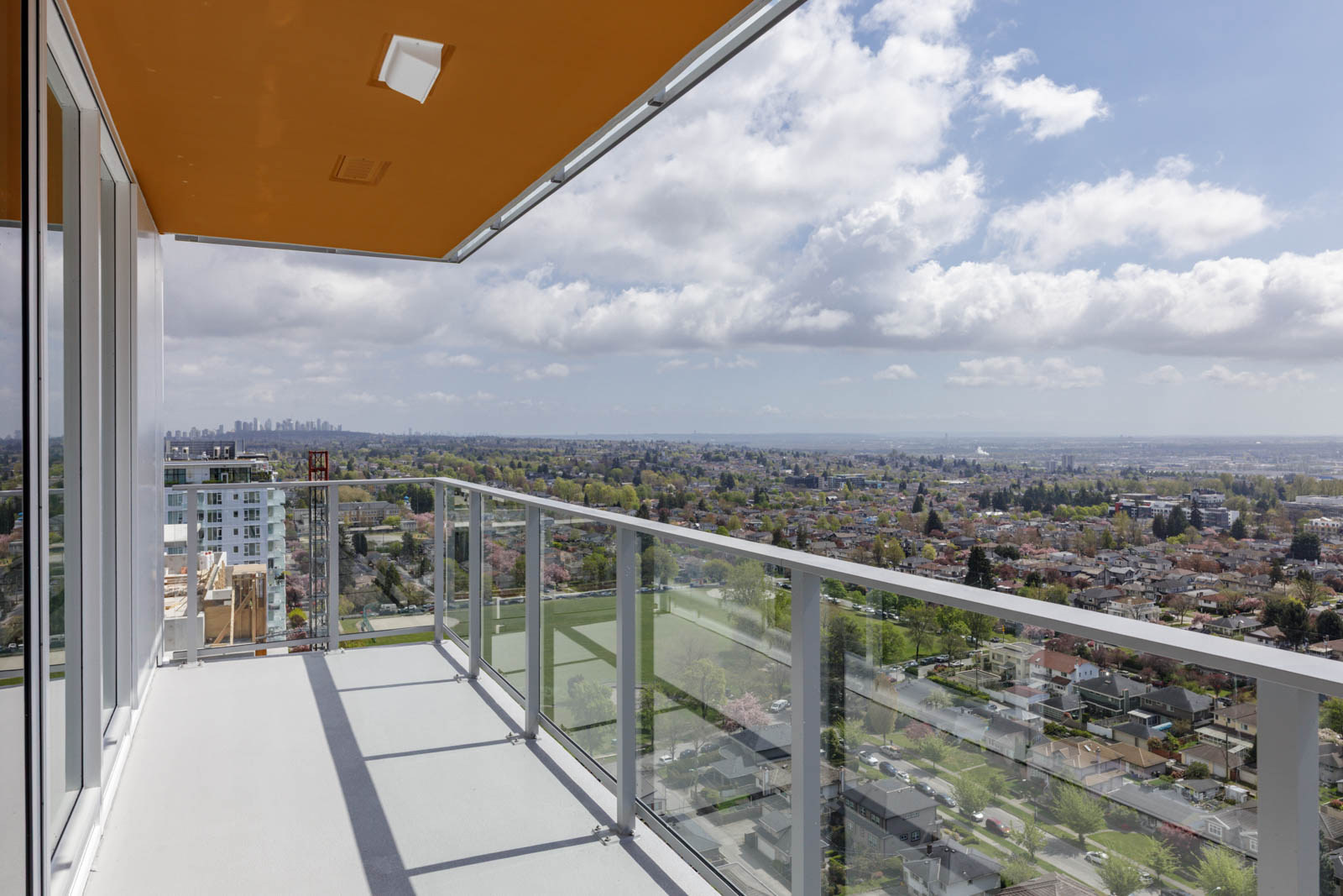 High-rise balcony with glass railing overlooking a cityscape, residential neighborhoods, and distant skyscrapers under a partly cloudy sky.