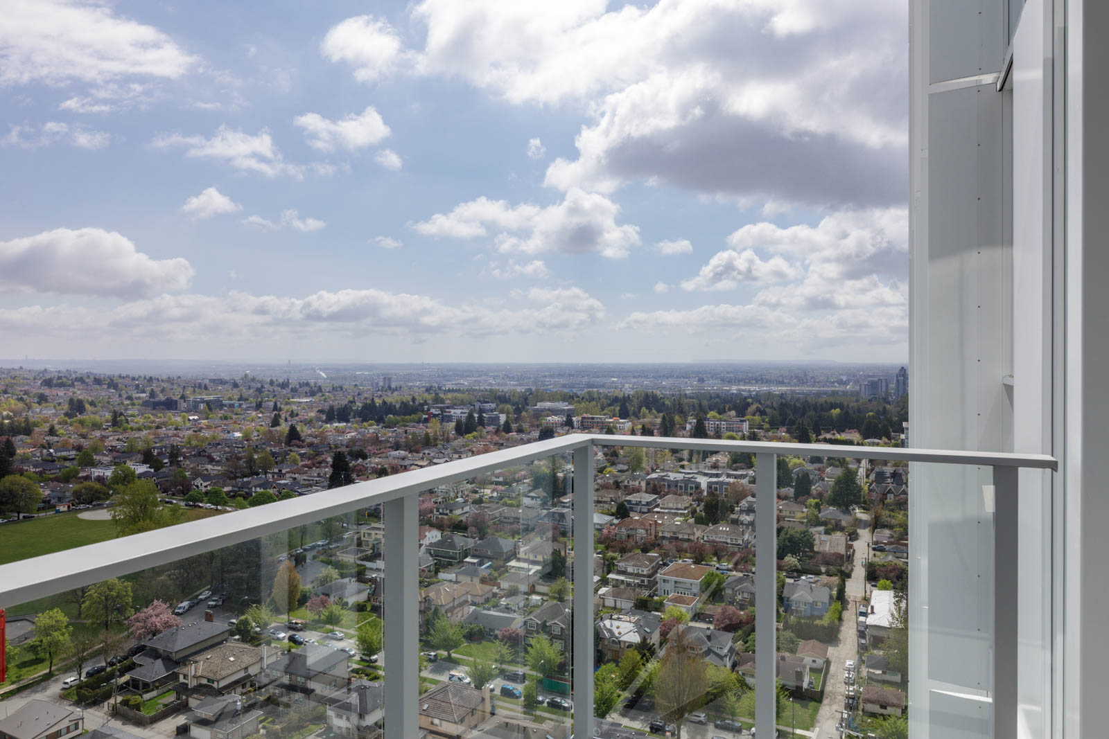 View from a high-rise balcony overlooking a suburban neighborhood with scattered clouds in the sky and distant cityscape on the horizon.