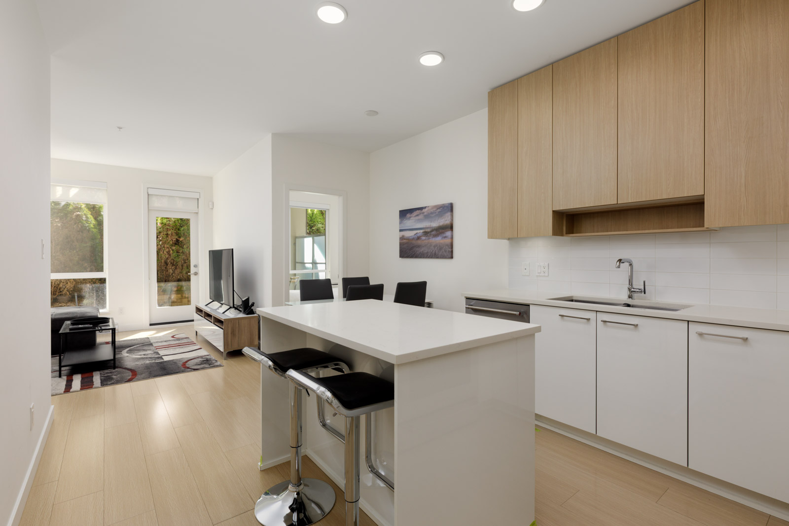Modern kitchen with white cabinets, wood accents, an island with two stools, and an open view into a bright living area with a TV and large windows.