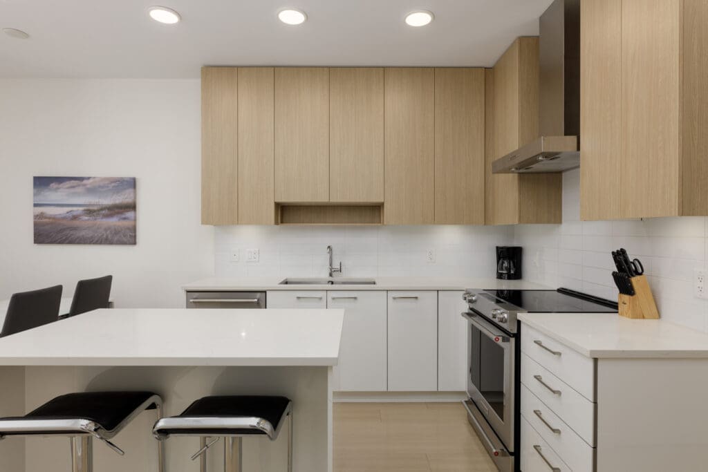 Modern kitchen with light wood cabinets, white countertops, stainless steel appliances, two black barstools, and a wall-mounted landscape photo.