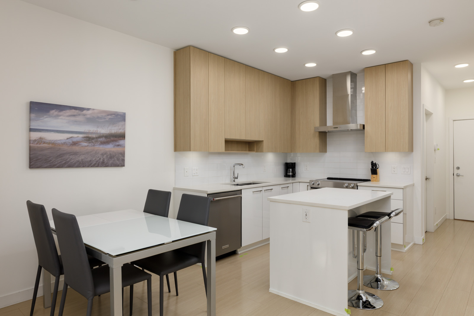 Modern kitchen with light wood cabinets, a small island with two stools, stainless steel appliances, and a dining table with four dark chairs. A landscape artwork hangs on the wall.