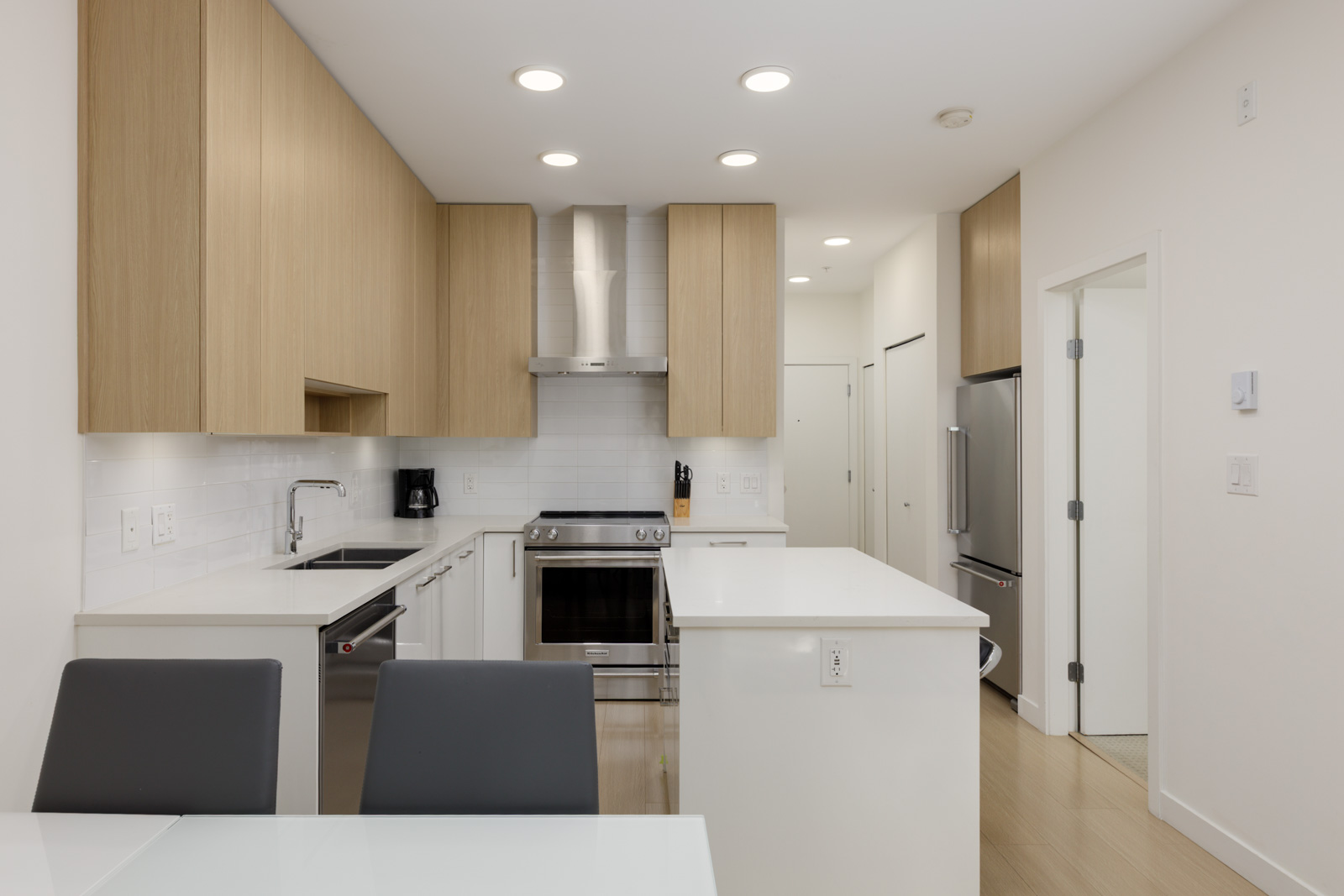 Modern kitchen with light wood cabinets, white countertops, stainless steel appliances, an island, and recessed lighting; dining table and chairs in the foreground.