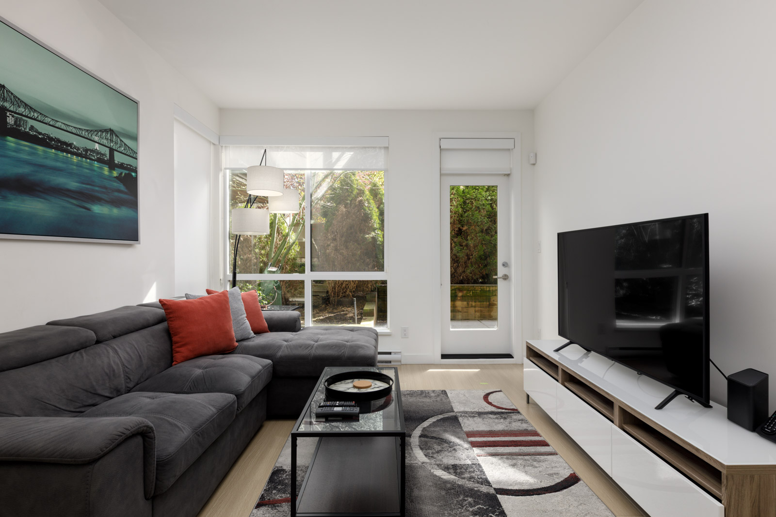 Modern living room with a gray sectional sofa, red accent pillows, a large flat-screen TV, artwork on the wall, and large windows letting in natural light.