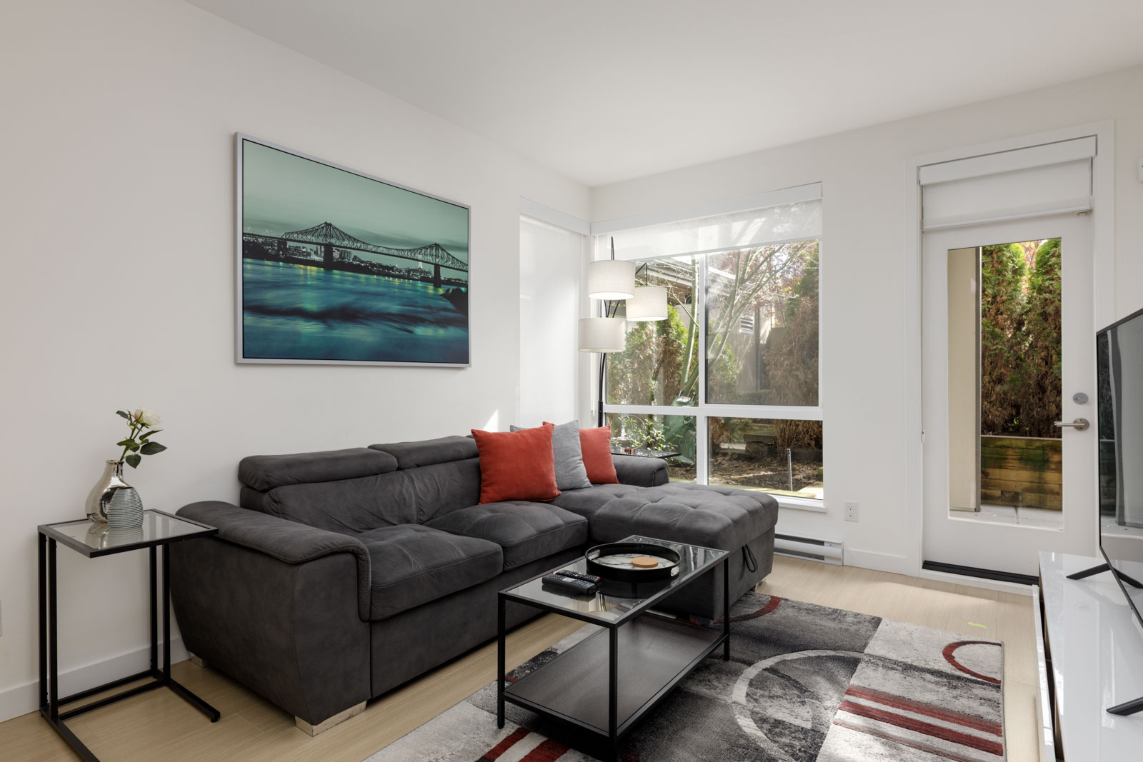 A modern living room with a gray sectional sofa, red accent pillows, a glass coffee table, large windows, and a framed bridge artwork on the wall.