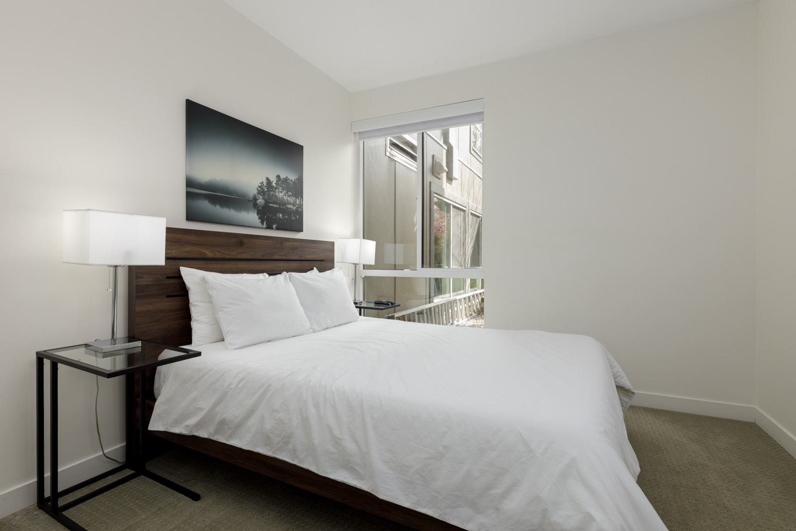 A modern bedroom with a wooden bed frame, white bedding, two table lamps, a side table, and a large window letting in natural light.
