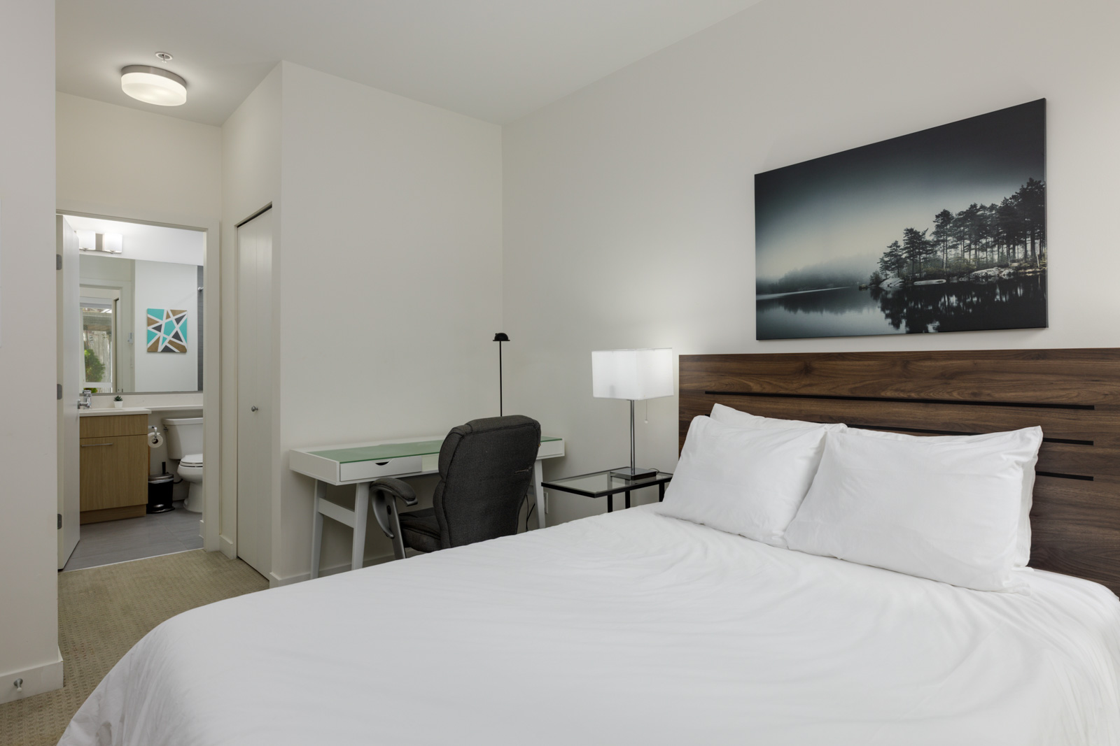A tidy bedroom with a made bed, white bedding, a wooden headboard, a desk with a chair, a lamp, and a bathroom visible in the background.