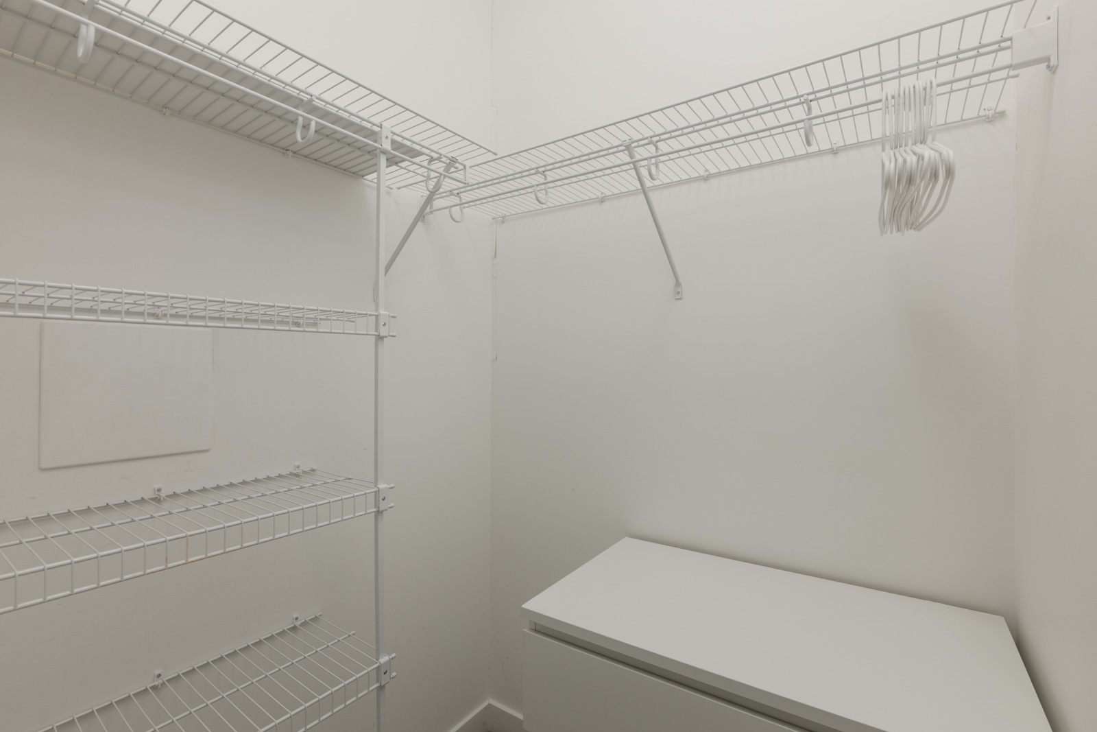 Empty walk-in closet with white wire shelves, a white cabinet, and a few empty white hangers on a top shelf against plain white walls.