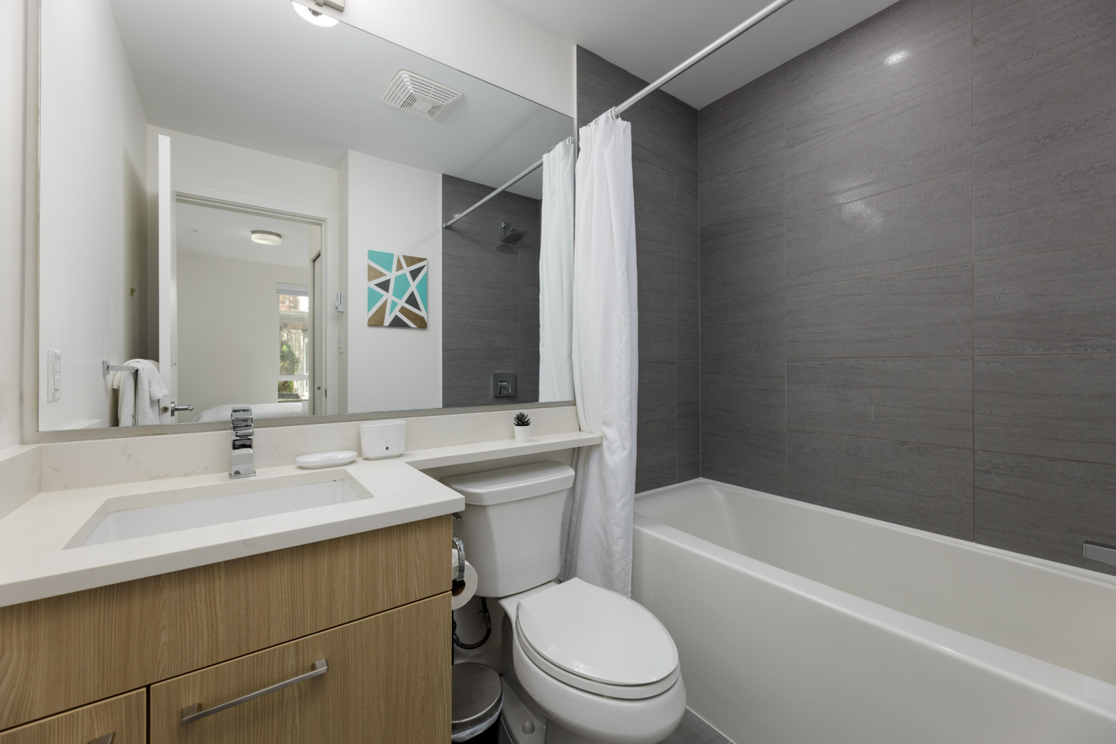 Modern bathroom with a light wood vanity, white sink, toilet, and a bathtub with a gray tiled wall and white shower curtain. Mirror and wall art are also visible.