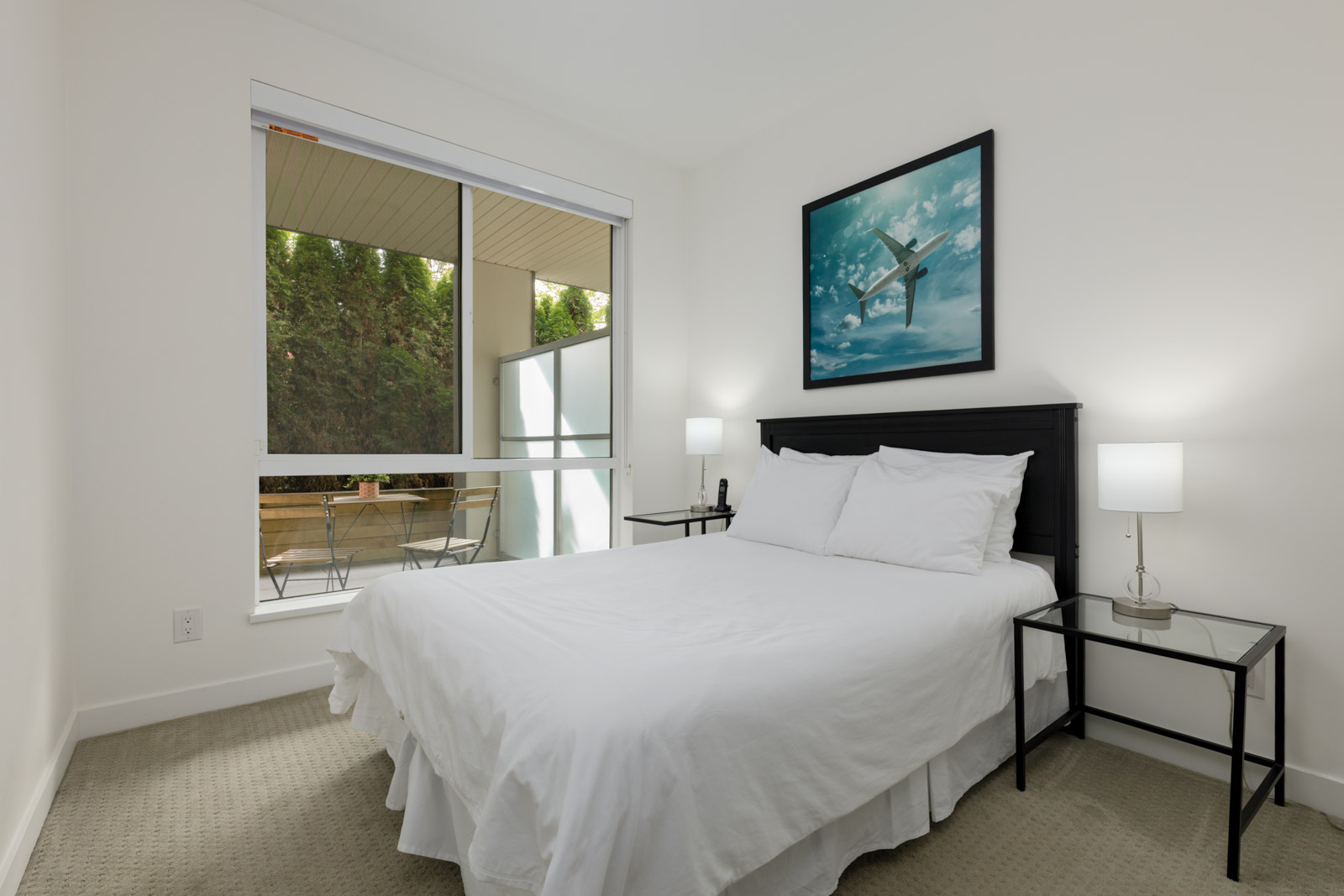 A bedroom with a neatly made bed, two bedside tables with lamps, a large window, and a framed airplane artwork above the headboard. A patio with chairs is visible outside.