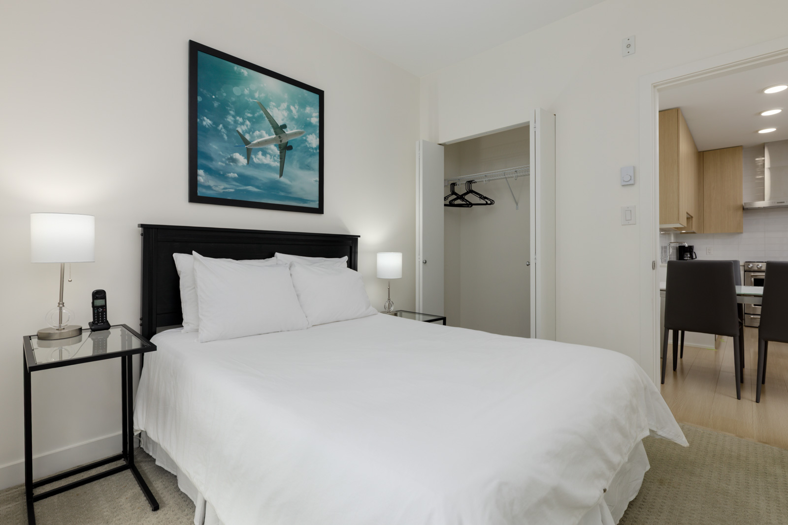 A neatly made bed with white linens, two bedside tables with lamps, an open closet, and wall art of an airplane; a kitchen is visible through an open door.