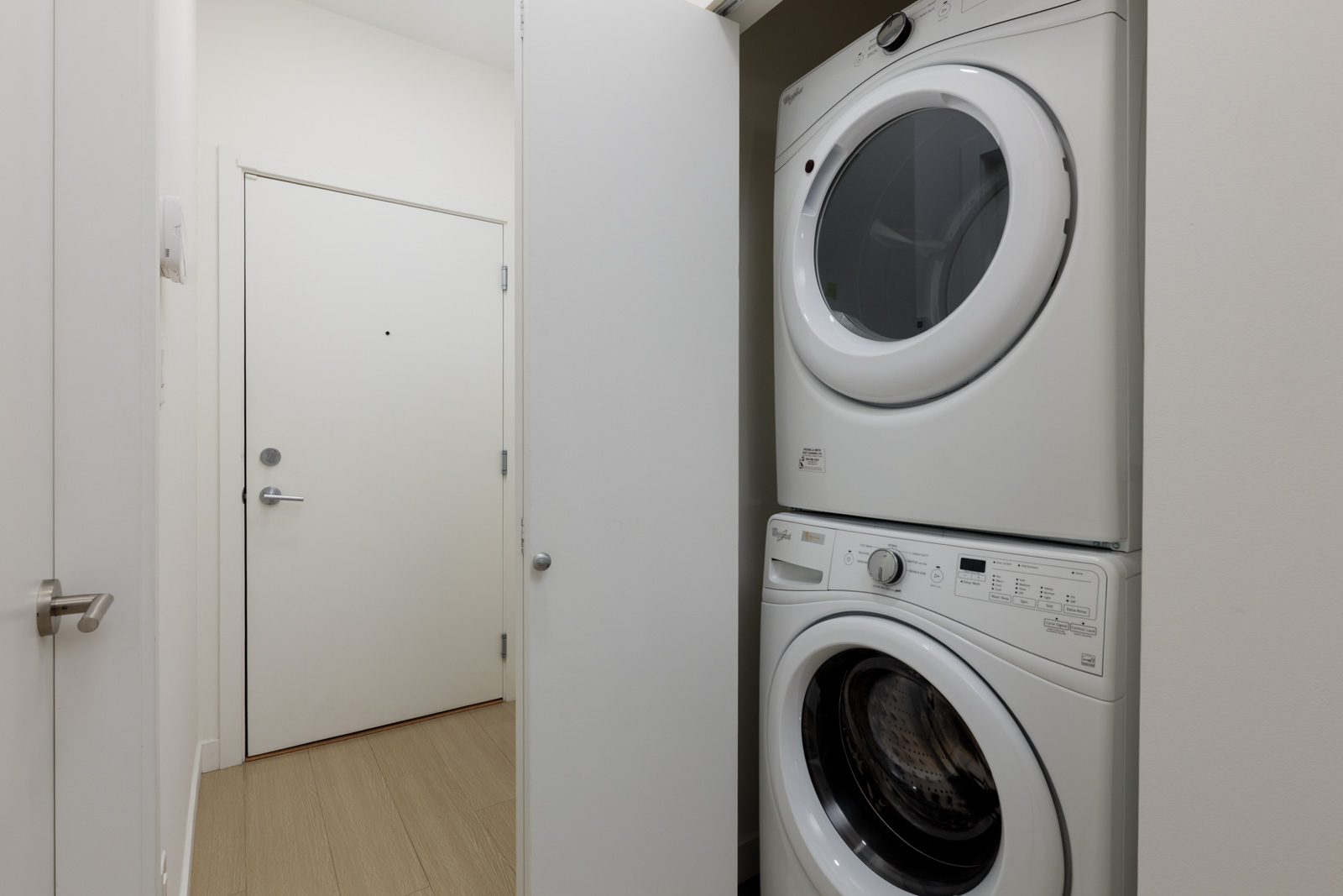 A stacked washer and dryer set is installed in a closet next to a closed entrance door in a minimalist, light-colored room.