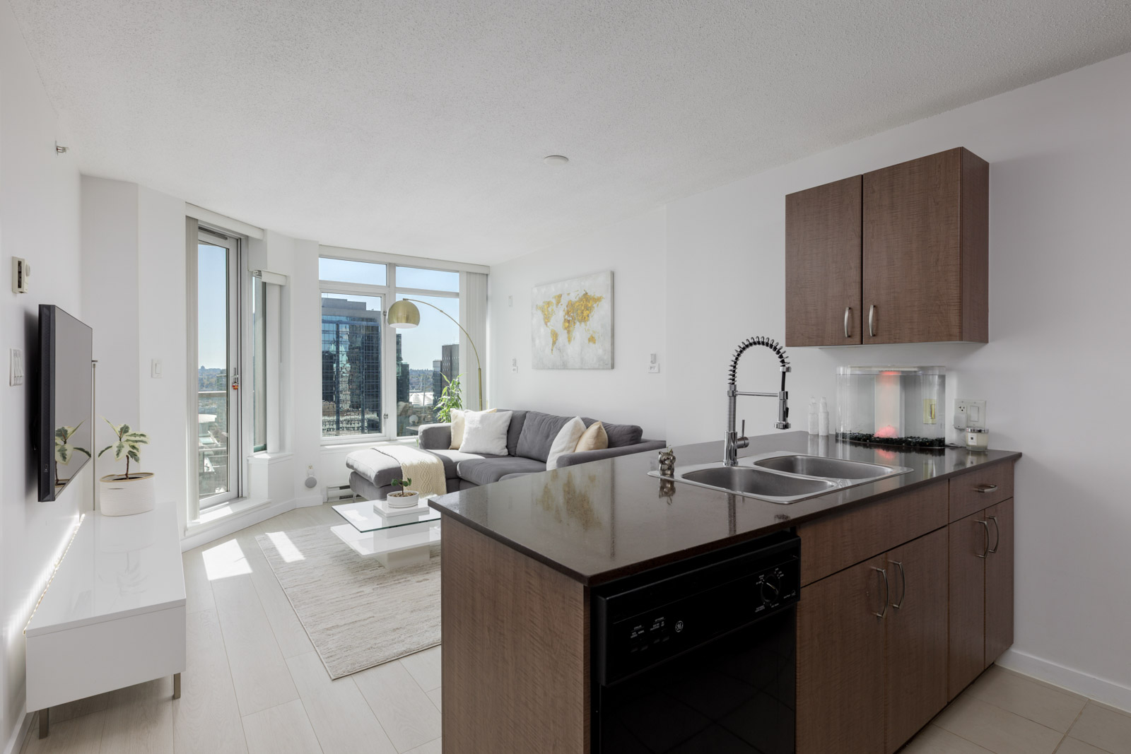 Modern apartment interior with a kitchen island, sink, living area, large windows, grey sofa, wall-mounted TV, and cityscape view.