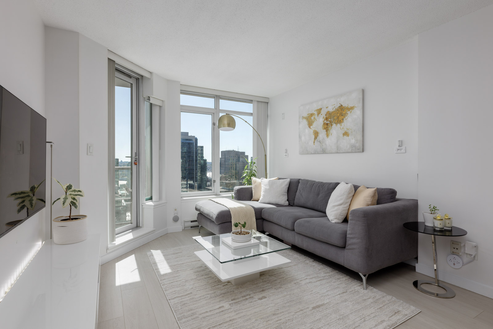 Modern living room with a gray sectional sofa, glass coffee table, large windows with city views, wall-mounted TV, and minimal decor including a world map art piece and potted plants.