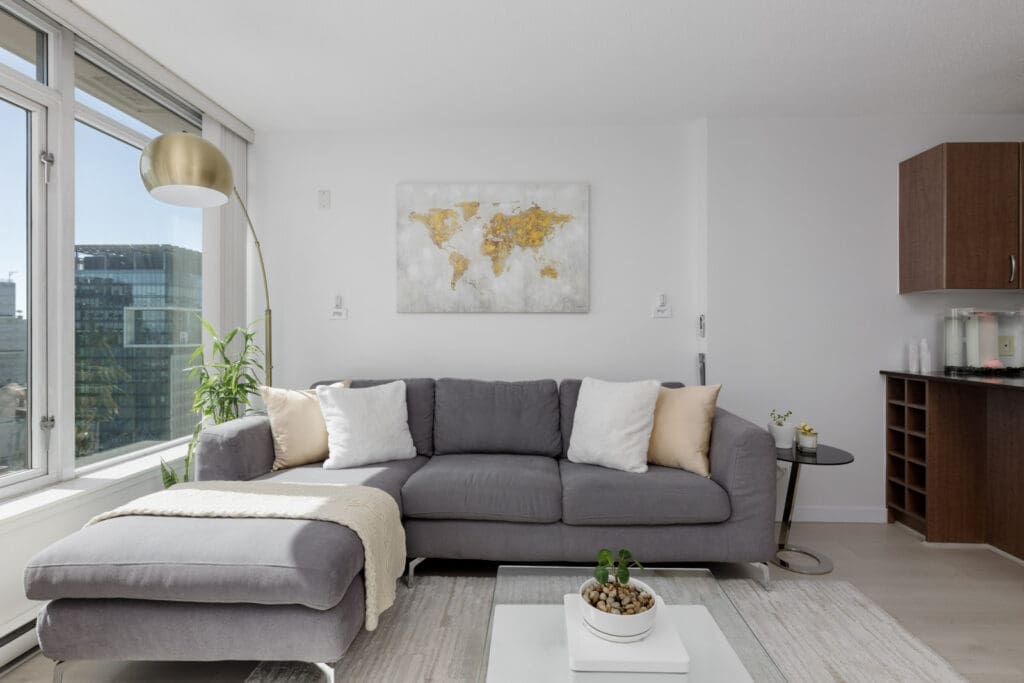 Modern living room with a gray sectional sofa, gold floor lamp, wall art of a world map, and large windows providing natural light.