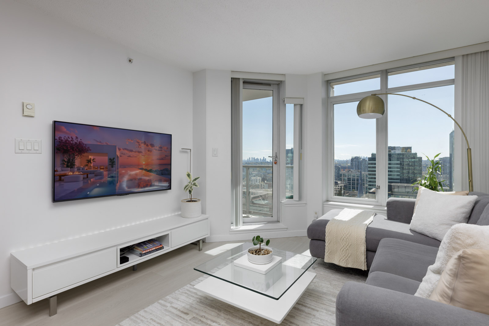 Modern living room with a gray sofa, glass coffee table, wall-mounted TV, large windows, and city skyline view.