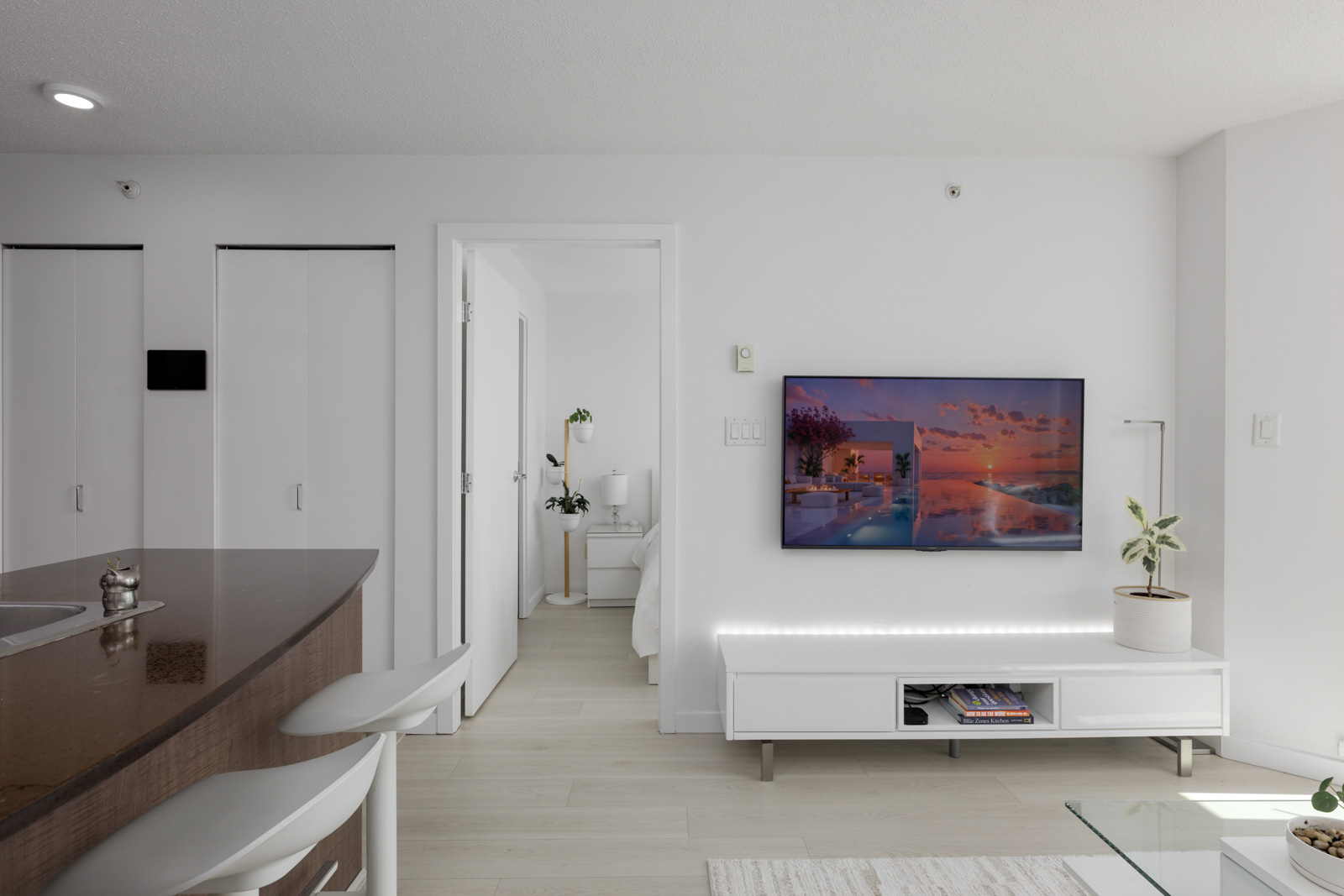 Modern living room with a wall-mounted TV, white media console, potted plant, and a view into a bedroom; kitchen counter with stools is visible on the left.