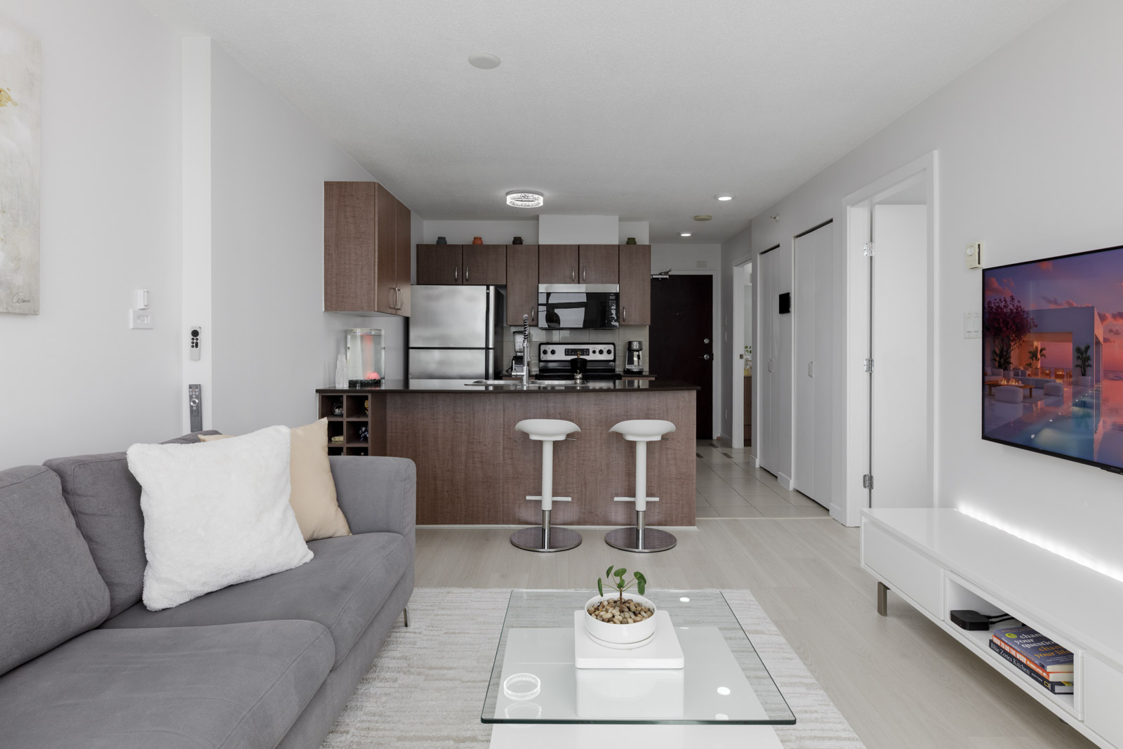 Modern apartment living room with a gray sofa, glass coffee table, and a kitchen in the background featuring stainless steel appliances and a breakfast bar with two stools.