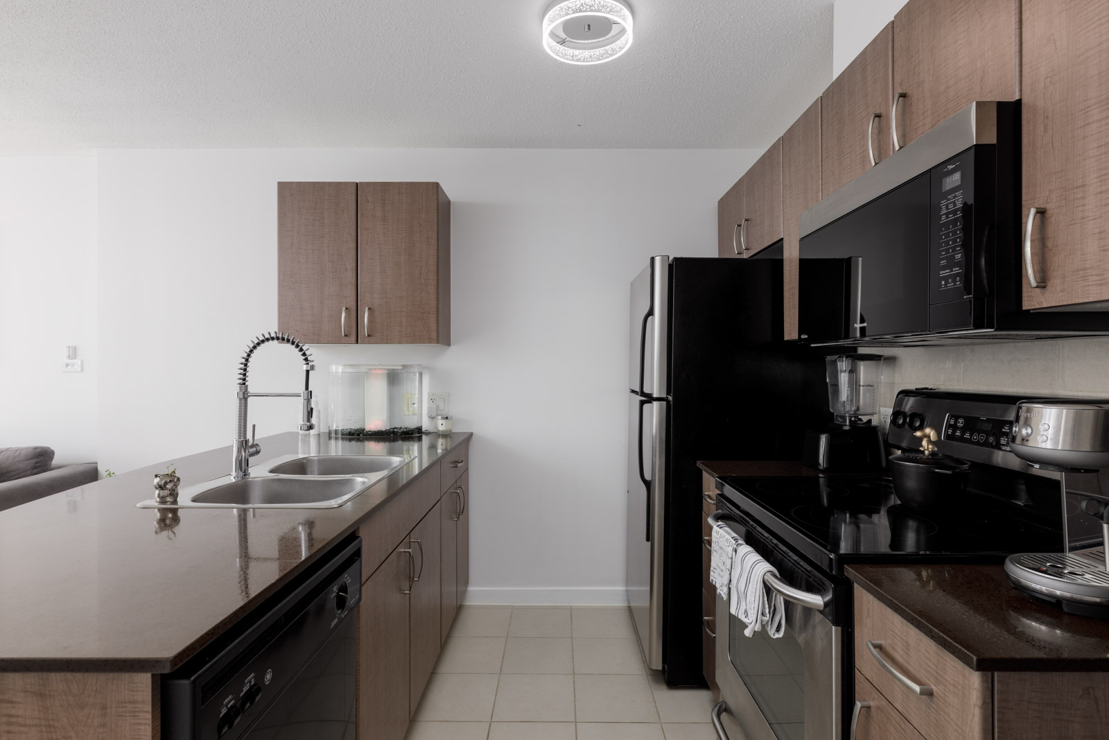 Modern kitchen with brown cabinets, black appliances, a double sink with a gooseneck faucet, and a dark countertop. A towel hangs on the oven door and a blender sits on the counter.