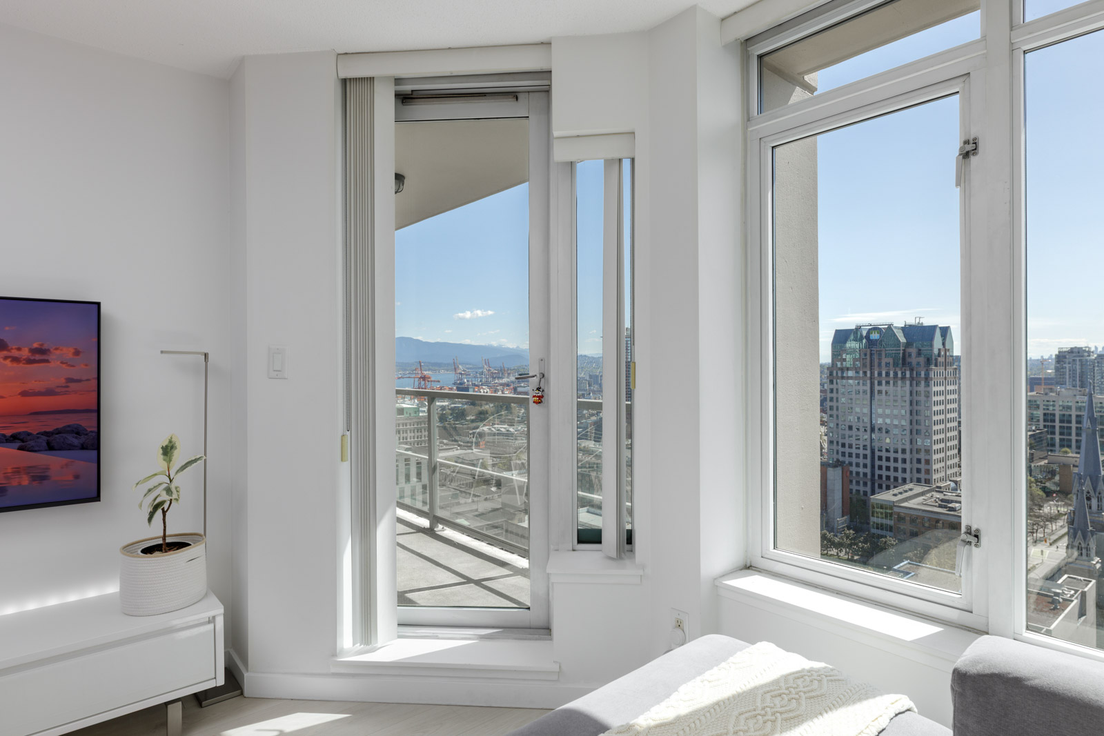 A modern high-rise apartment room with large windows, a balcony door open to a city view, and a small plant on a white cabinet.