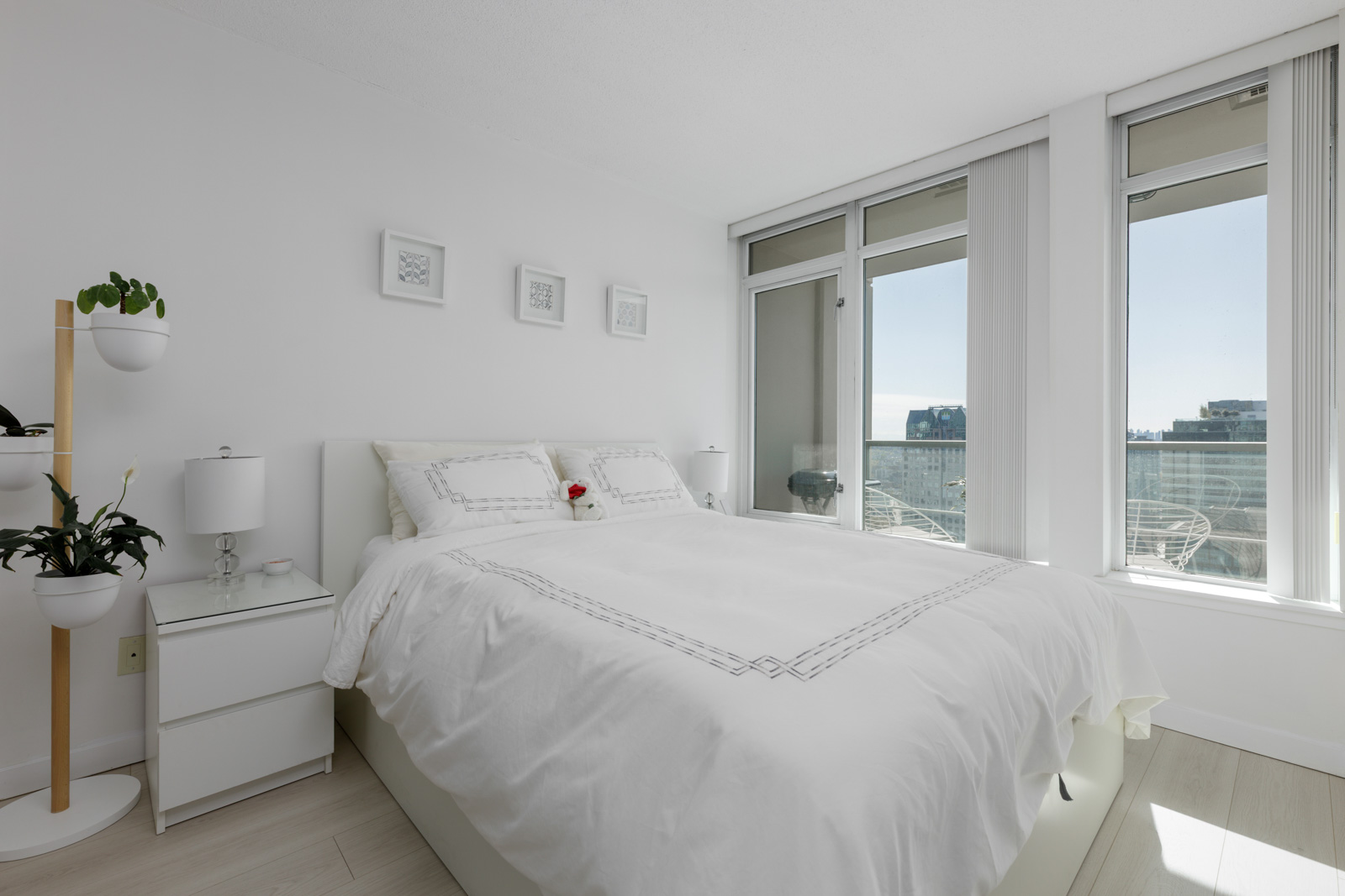 A modern, bright bedroom with a white bed, two nightstands, potted plants, and large windows overlooking a balcony and cityscape.