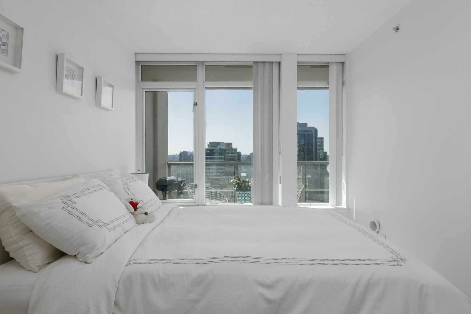 A modern bedroom with a white bed and bedding, three framed pictures on the wall, and large windows overlooking a balcony and city buildings.