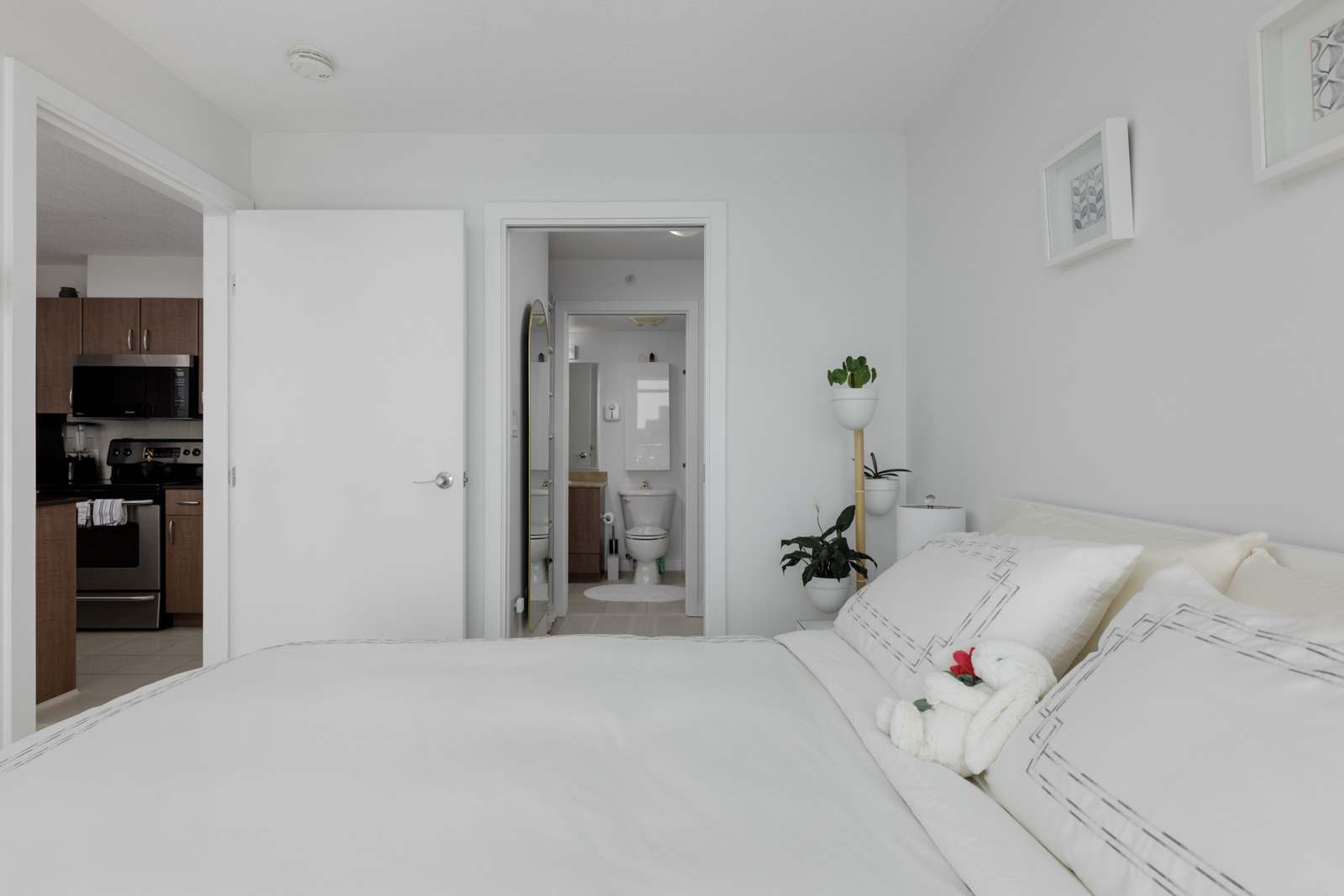A modern bedroom with a neatly made bed, white linens, and plants, showing a view into an en-suite bathroom and a kitchen area in the background.