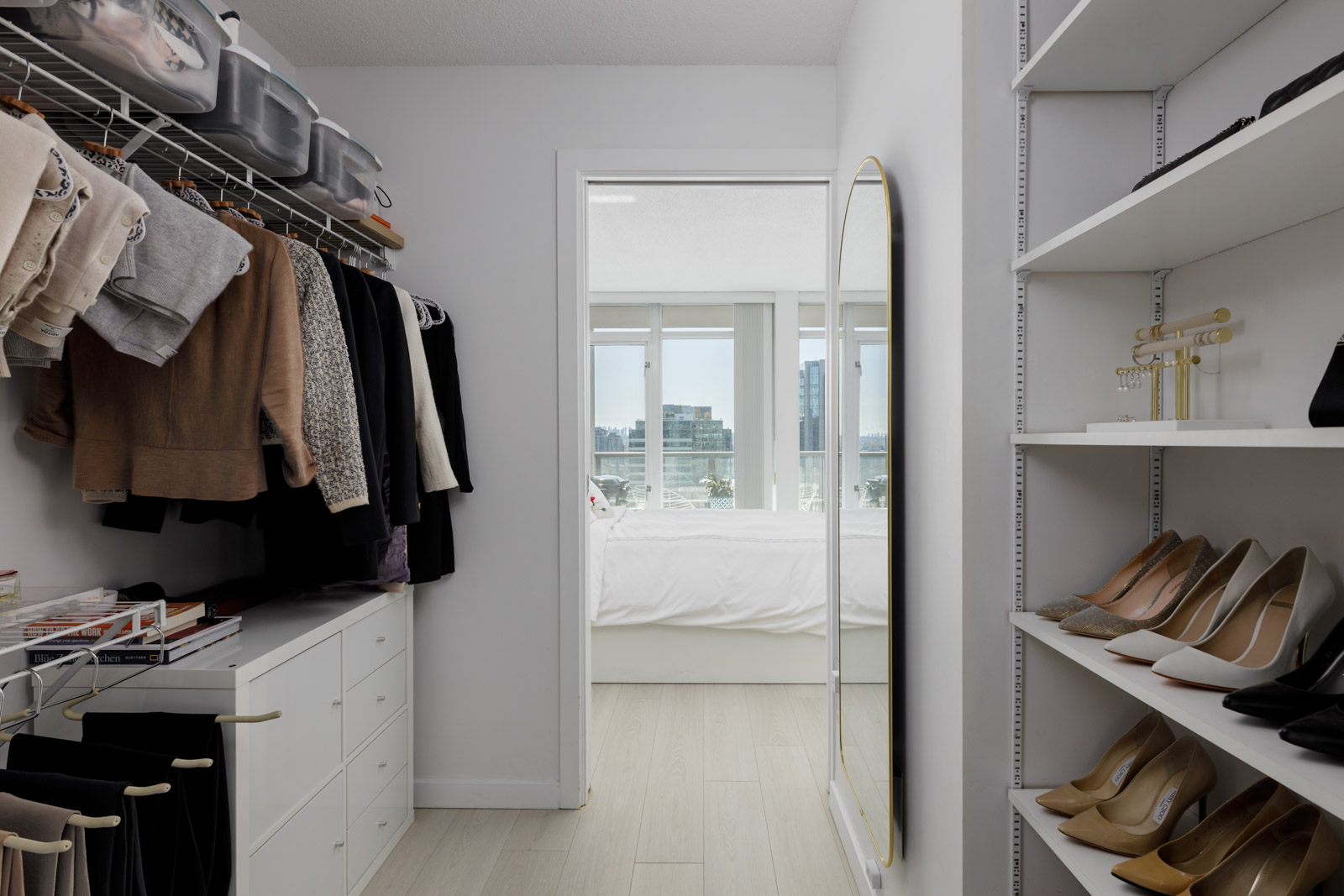 A modern walk-in closet with neatly organized clothes, shoes, and accessories, leading to a bright bedroom with large windows and a city view.