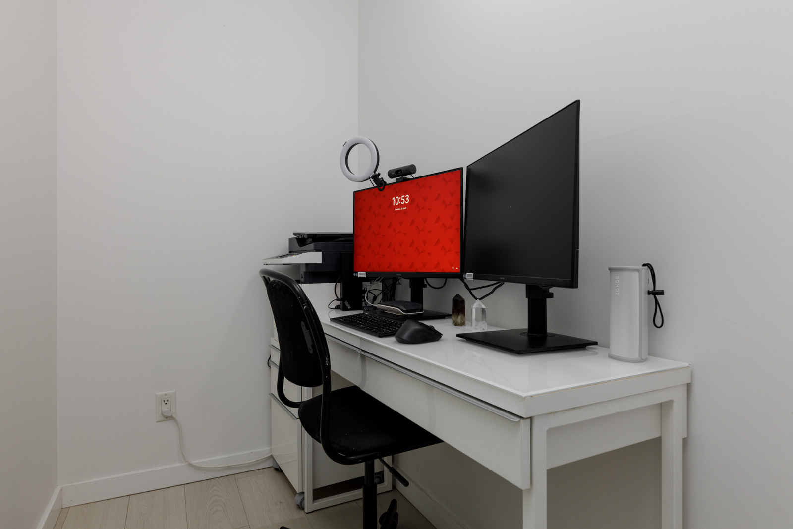 A minimalist home office with a white desk, two monitors, a keyboard, a webcam, a ring light, a black chair, and a printer against a plain white wall.