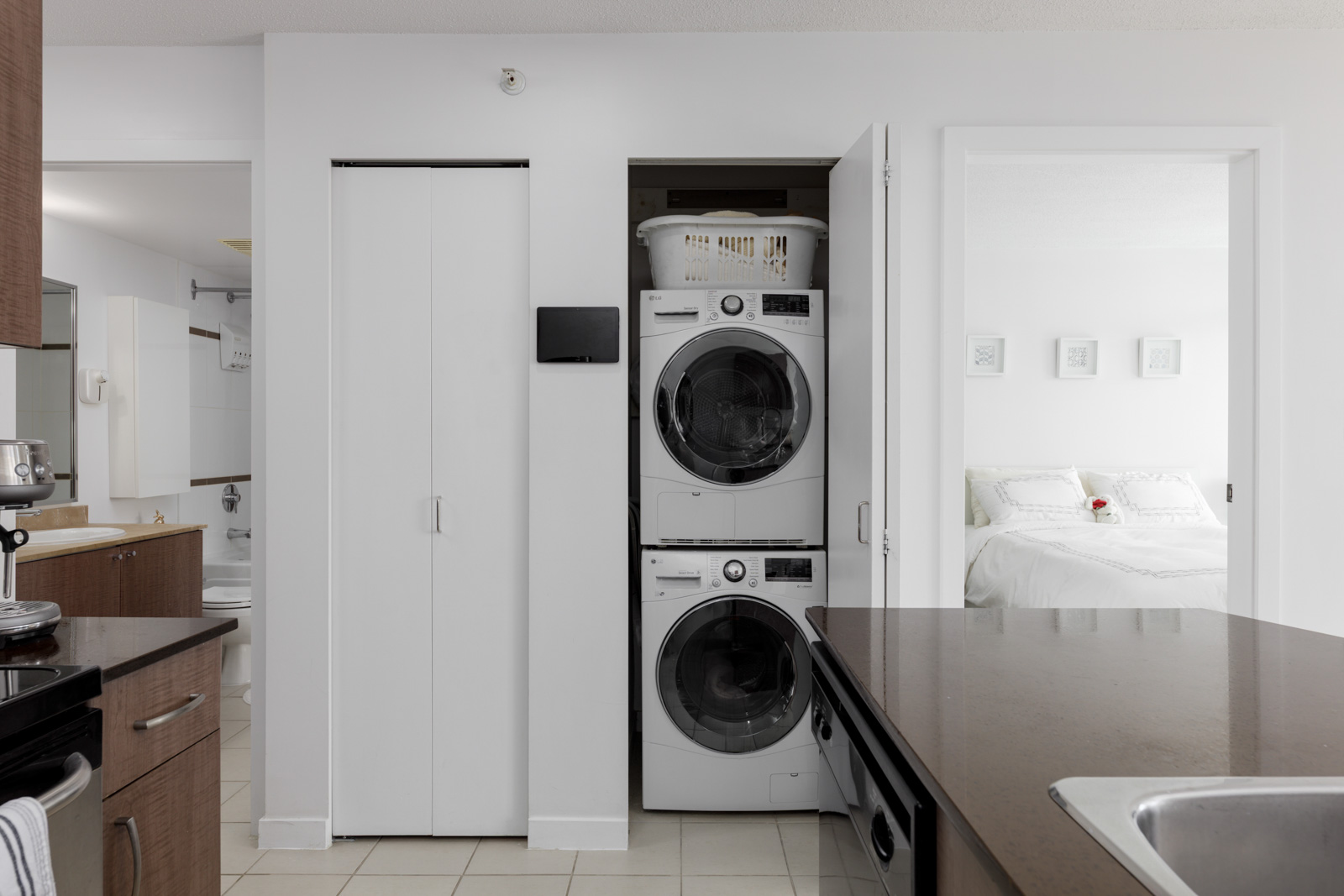 A stacked washer and dryer in a closet between a bathroom and a bedroom in a modern, white apartment interior.
