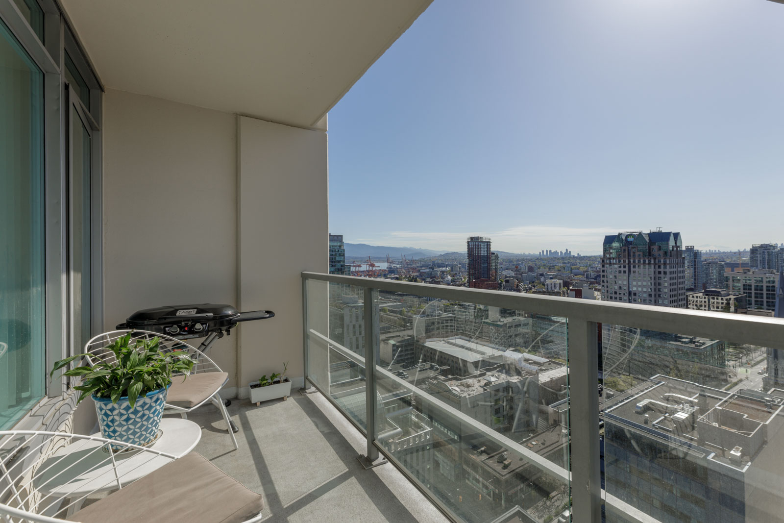 High-rise balcony with two chairs, a small table with a potted plant, and a barbecue grill, overlooking a cityscape under a clear sky.