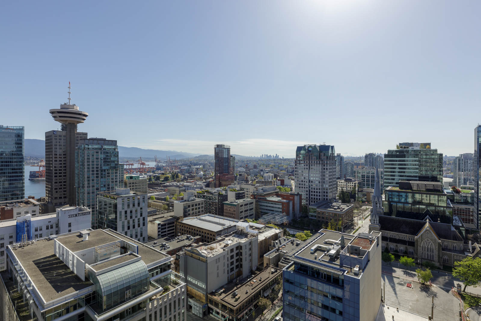 Aerial view of downtown Vancouver with office buildings, the Harbour Centre, and mountains visible in the distance under a clear sky.