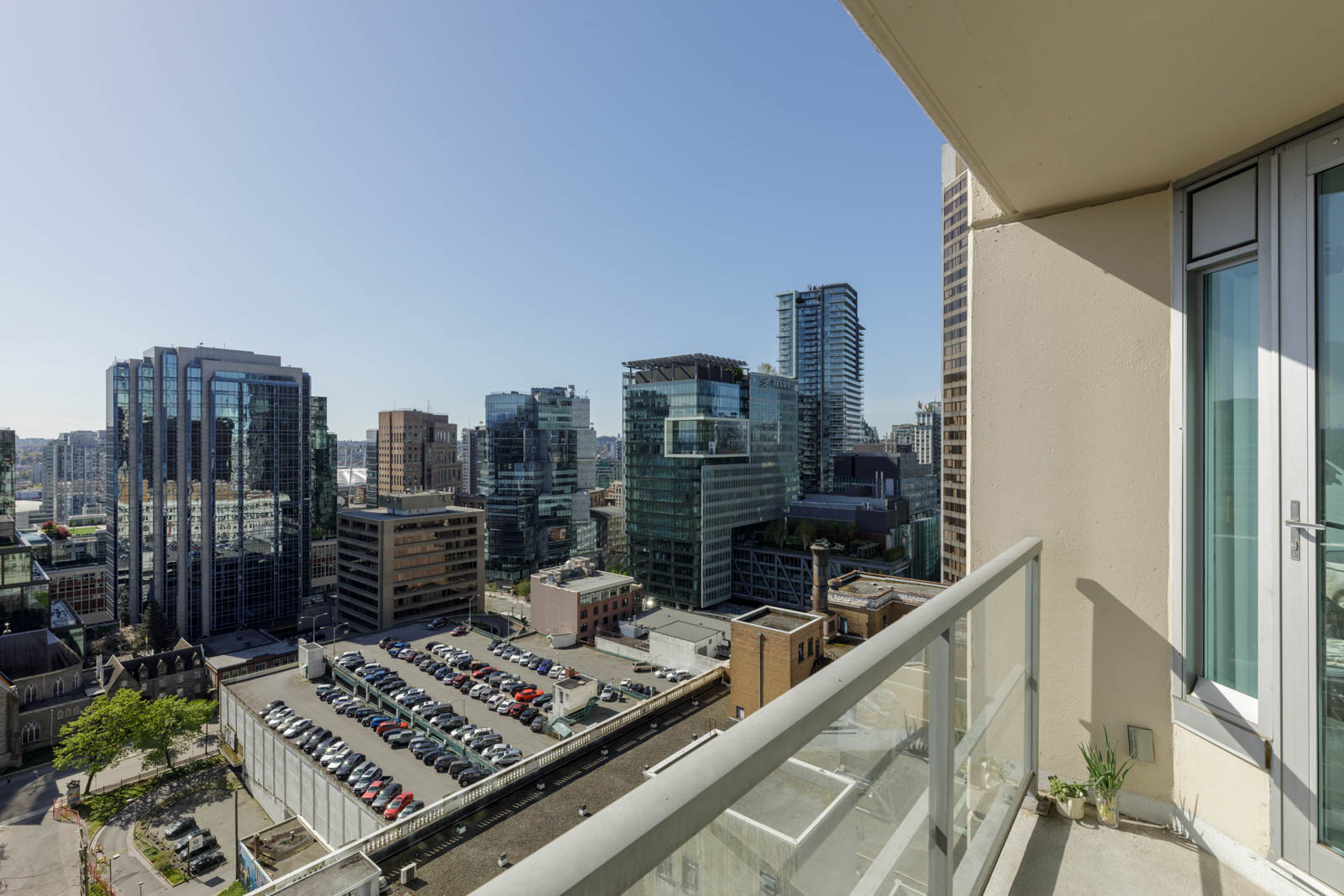 View from a balcony showing a cityscape with modern high-rise buildings, a parking lot below, and clear sky in the background.